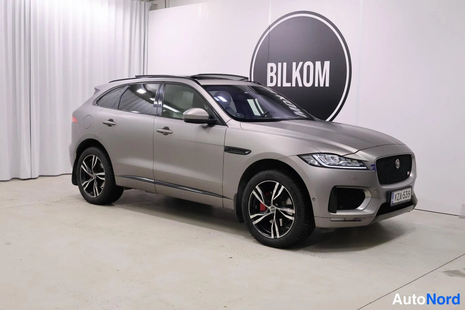Etuviisto, nokka oikealle: Harmaanbeige Jaguar F-Pace etuokoliskolta oikealle, punaiset jarrusatulat ja -kilpi näkyvissä.