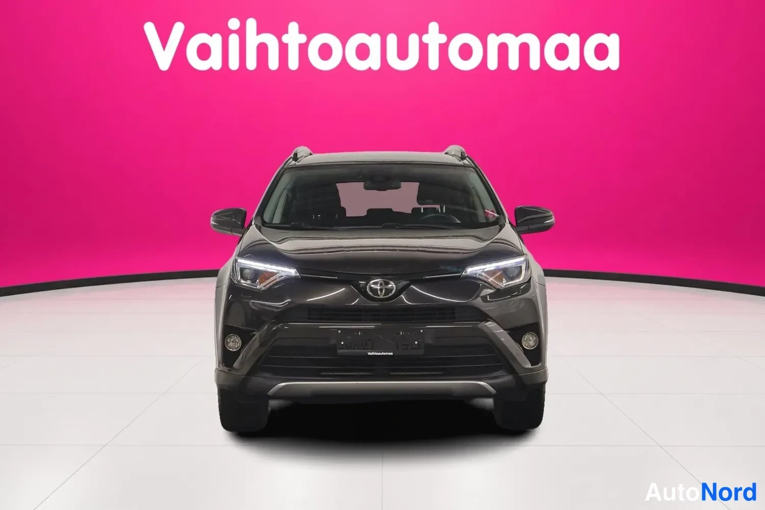 Edestä: Mustanruskean Toyota RAV4:n etunäkymä, Toyota-merkki ja etumaski ilmeikkäällä vaaleanpunaisella taustalla.