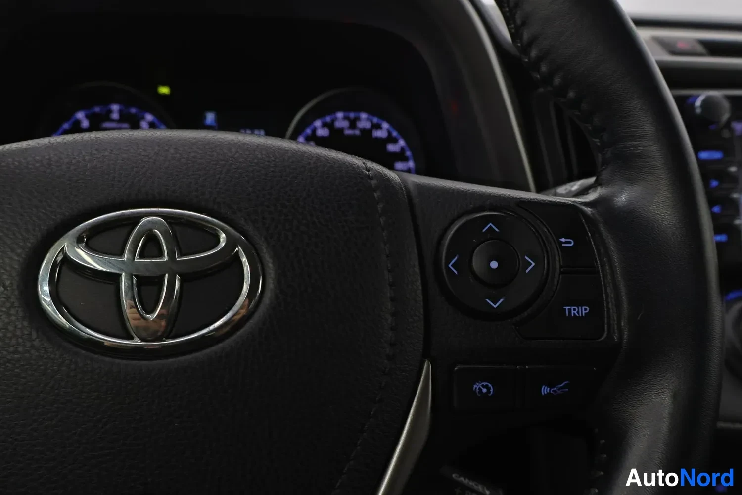 Yksityiskohta: Toyota-merkillä varustettu ohjauspyörä lähikuvassa, näkyvissä myös rattiin integroidut napit ja taustalla mittariston näytöt.