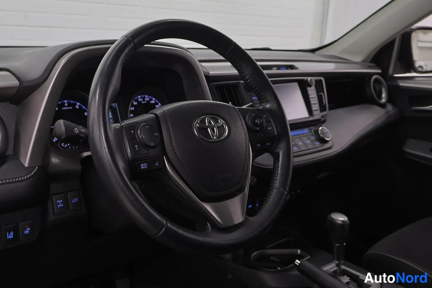 Muu: Ohjauspyörän lähikuva Toyota-merkillä, mittaristo ja keskikonsolin infotainment-näyttö näkyvät taustalla.