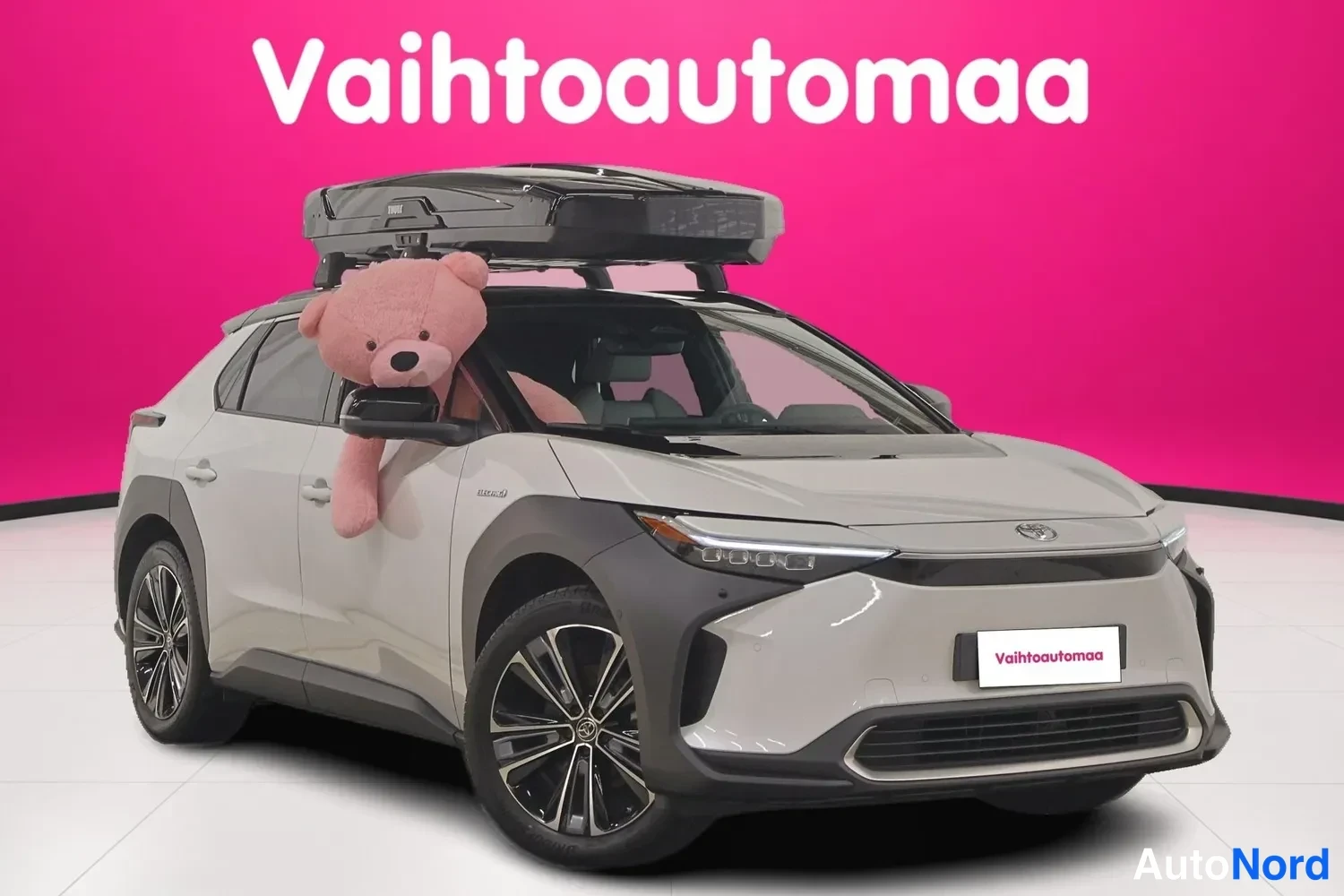 Etuviisto, nokka oikealle: Toyota bZ4X, valkoinen, etuosa 3/4 oikealta, kattolekella, taustana Vahtoautomaan vaaleanpunainen studio.