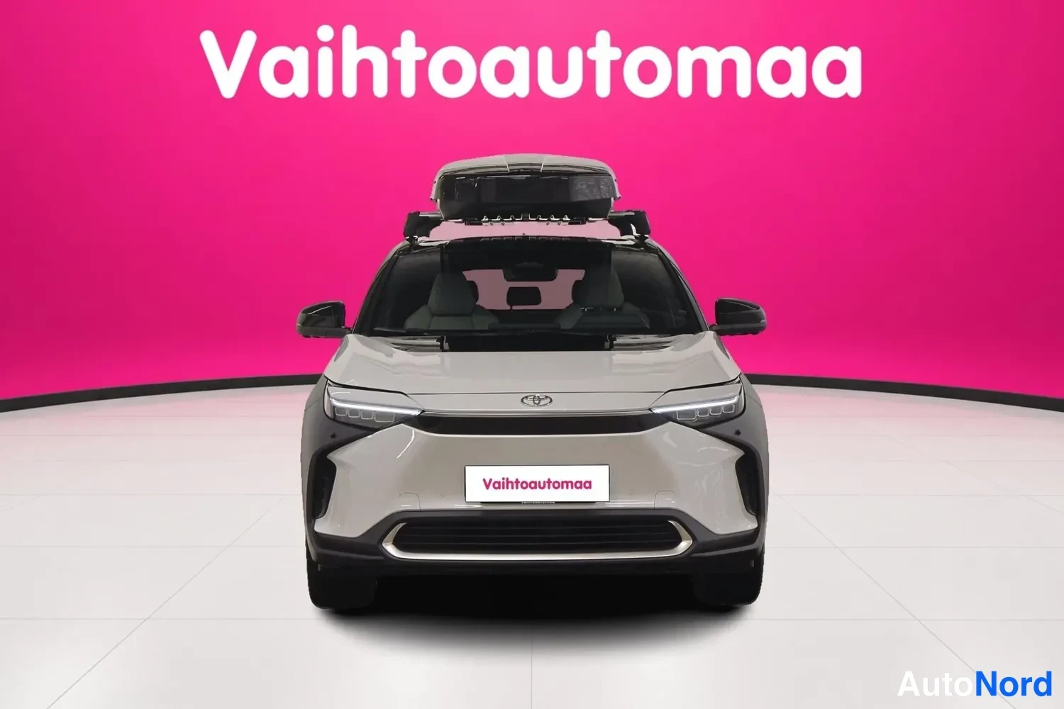 Edestä: Toyota bZ4X, valkoinen, suora etukuva, kattotelineellä katti-iboxi, tausta vaaleanpunainen studio.