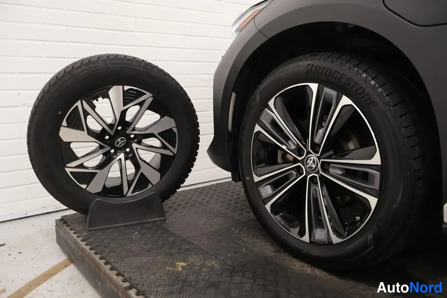 Yksityiskohta: Kaksi Toyota-kevytmetallivannetta paksurengaskumilla; vasemmalla irrotettu rengas telineessä, oikealla rengas auton pyörässä.