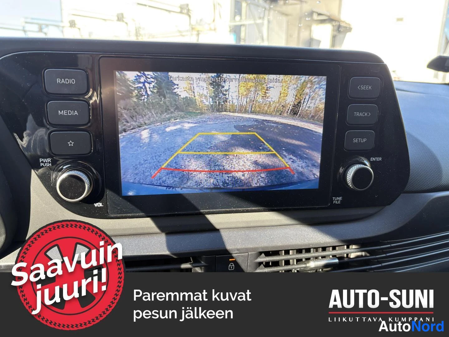 Muu: Infotainment-näyttö näyttää peruutuskameran kuvaa parkkitolinjoilla; ylälaidassa teksti 'Tarkista ympäristö turvallisuutesi takia'.