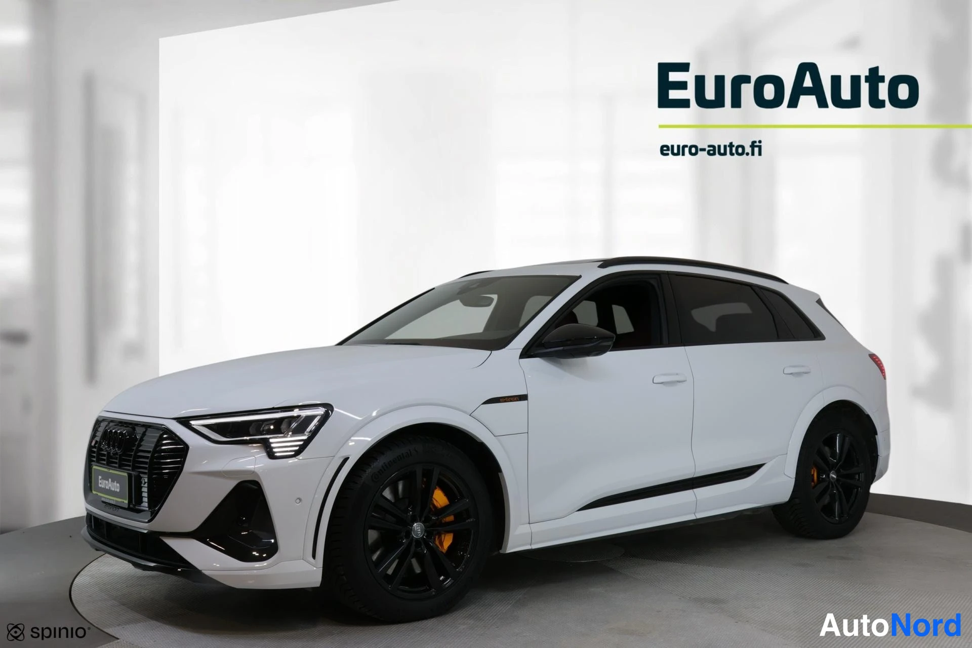 Etuviisto, nokka vasemmalle: Valkoinen Audi e-tron etukolmanneskuvassa, keula kuvan vasemmalla laidalla; musta etusäleikkö, mustat vanteet ja oranssit jarrusatulat.