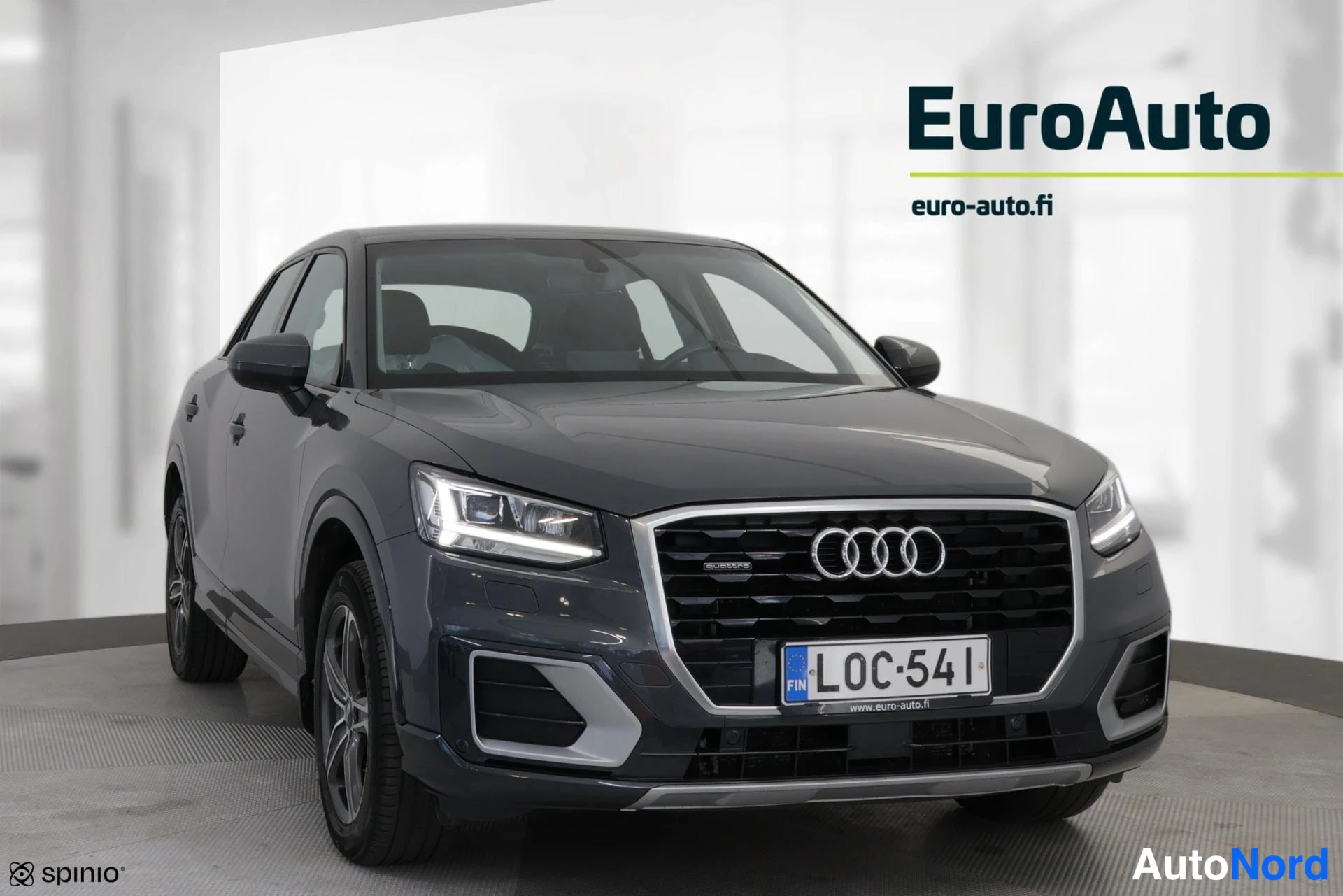 Edestä: Viisivärinen harmaa Audi Q2 suoraan edestäpäin. Etusäleikkö Audi-logolla, LED-lamput ja rekisterinumero ovat selkeästi näkyvissä valkoisessa studioympäristössä.