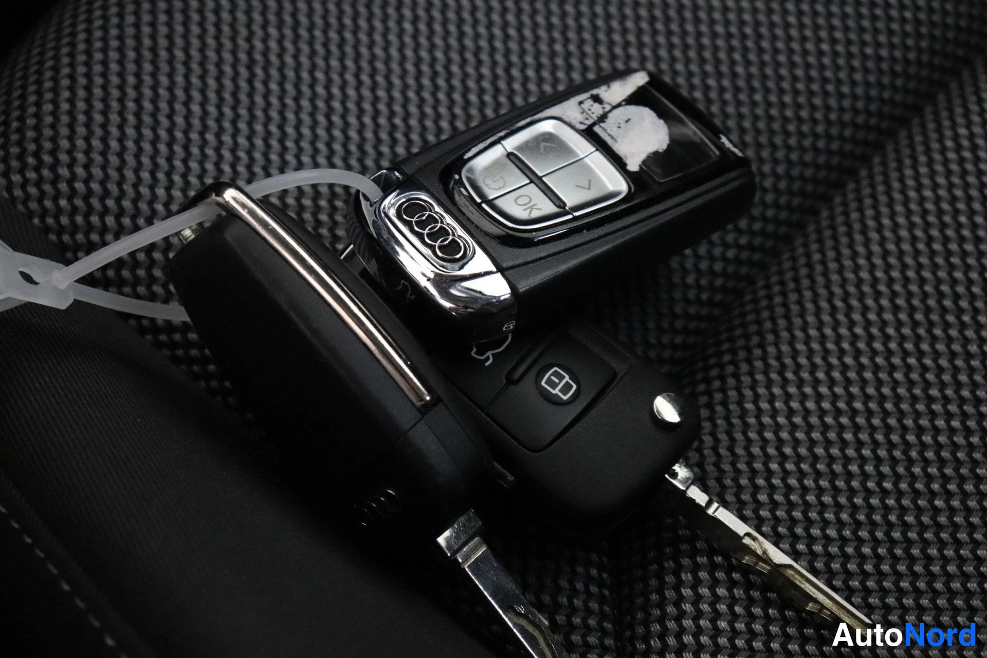 Muu: Kaksi auton avainta tekstiiliä vasten: Audi-logo varustettu radiokauko-ohjain ja perinteinen avain.