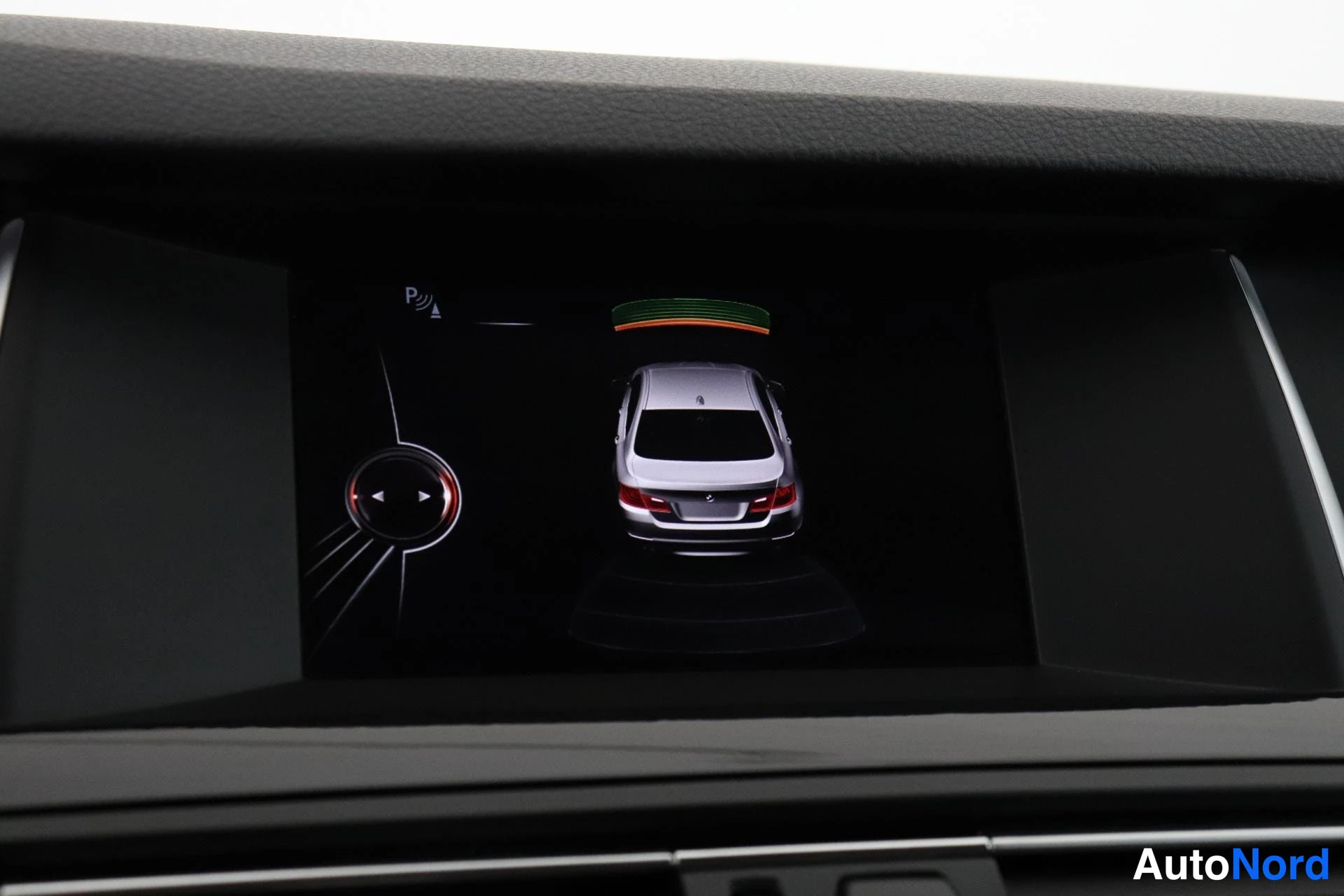 Yksityiskohta: Infotainment screen displaying a rear-view camera perspective of a silver car with parking sensor graphics and navigation controls on the left side of the displ