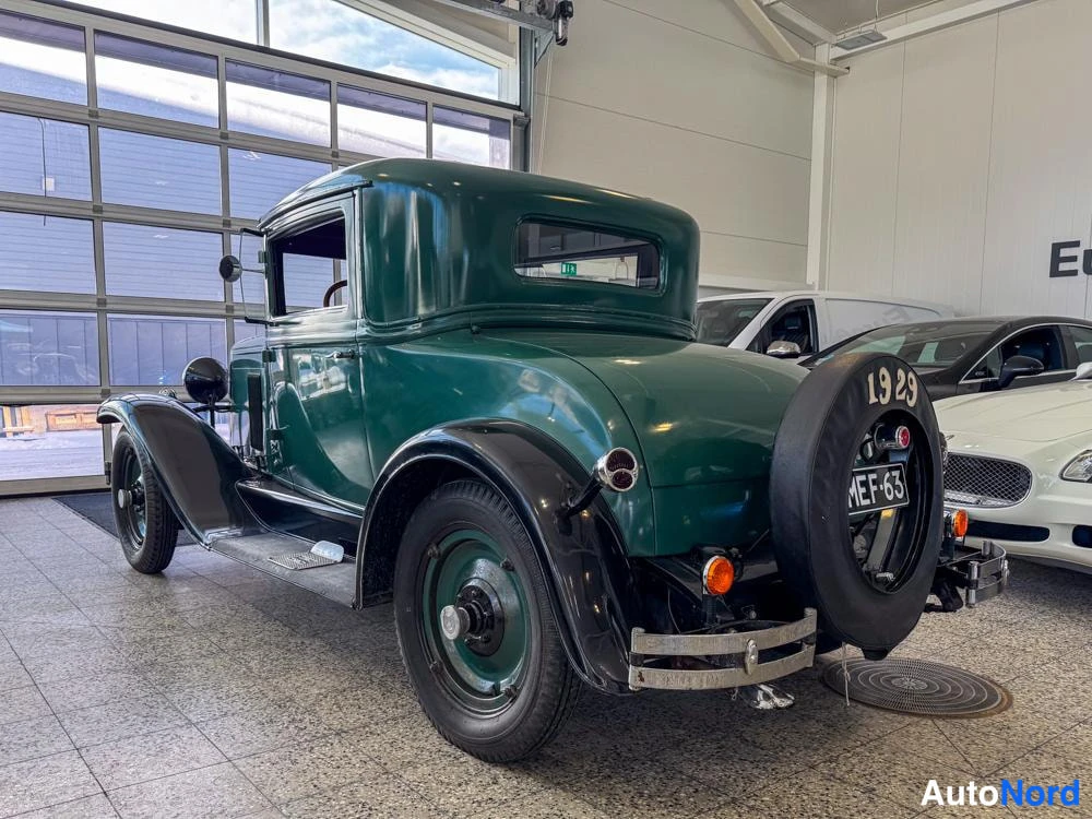 Takaviisto: Vihreä vintage-kupevaunu takakulmasta nähtynä: varapyörä tekstillä 1929 ja rekisterikilpi, perän ja vasemman kyljen näkyessä valoisassa autonhallissa.