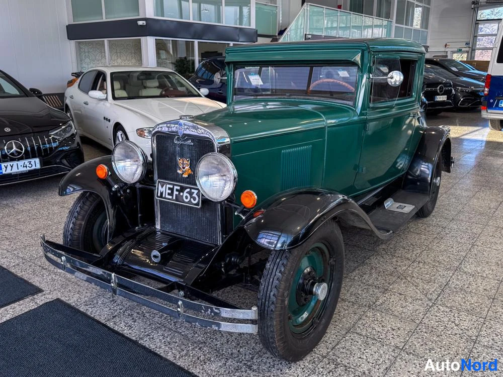 Etuviisto, nokka vasemmalle: Front three-quarter view of a vintage green coupé with black running boards and fenders inside a showroom. The front face (grille, headlights) and the