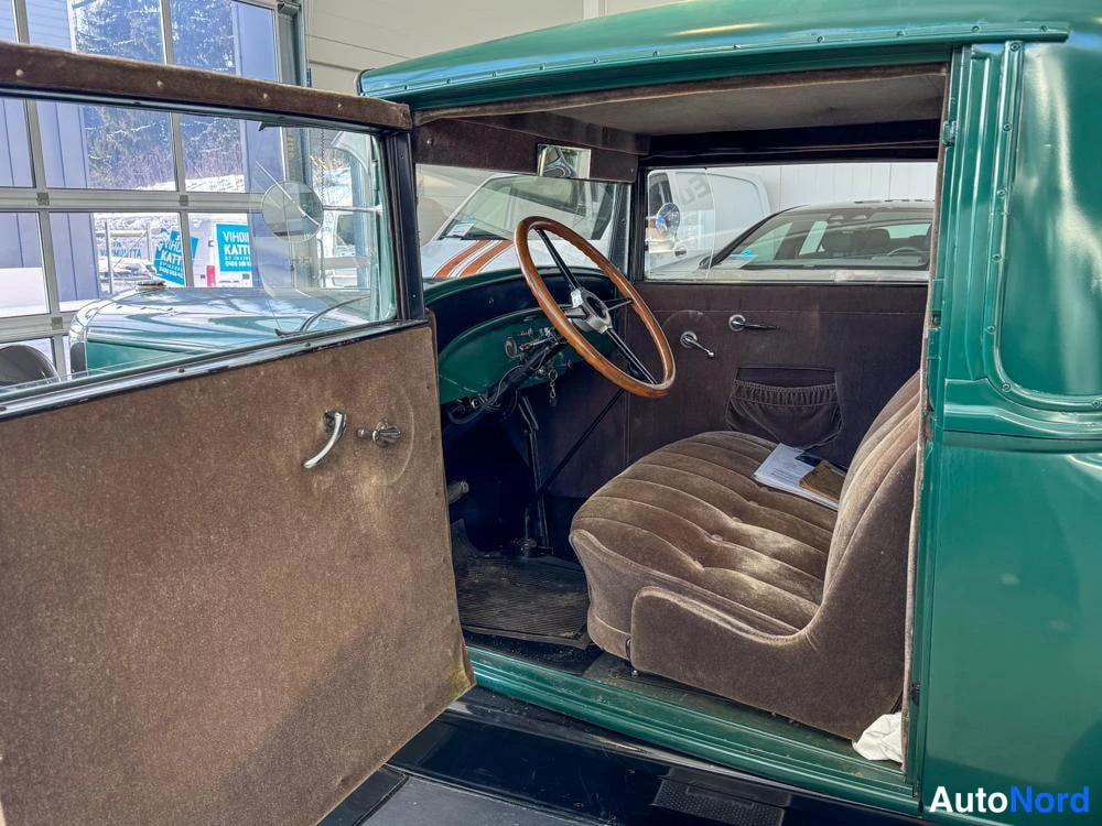 Ohjaamo: Interior shot showing the driver's side with the door open. A brown bench seat spans the width of the cabin next to a large wooden steering wheel.