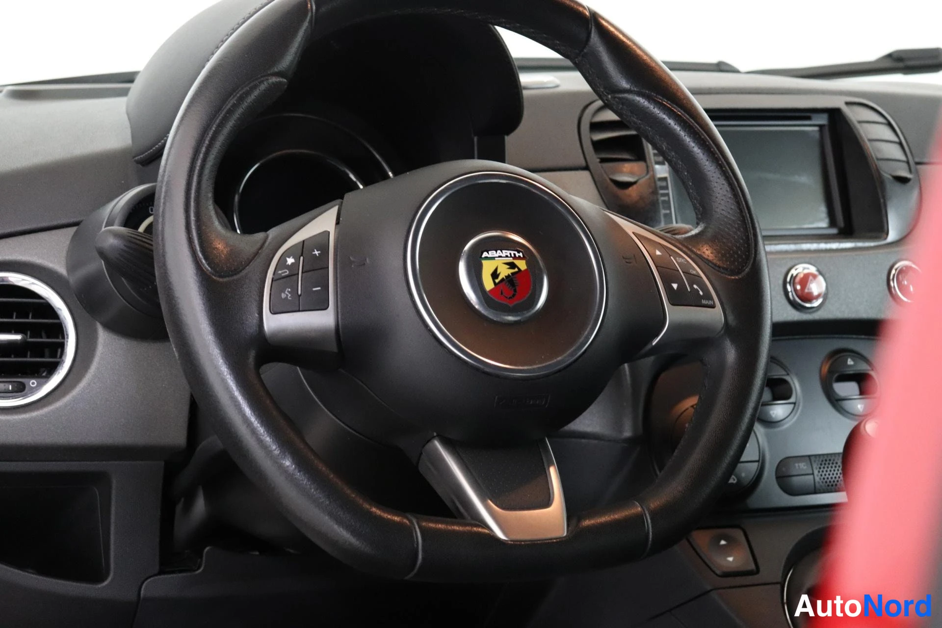 Yksityiskohta: Close-up of the black leather-wrapped steering wheel with silver multifunction controls on the right spoke and the red-start button visible on the dashboard to