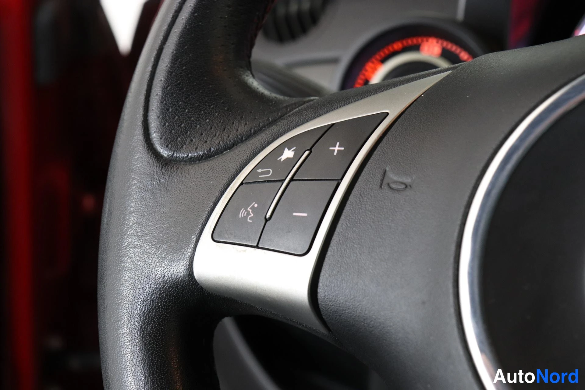 Yksityiskohta: Extreme close-up detail of the steering wheel control cluster showing buttons for volume control (+/-), track skip (arrows), mute and audio source selection.