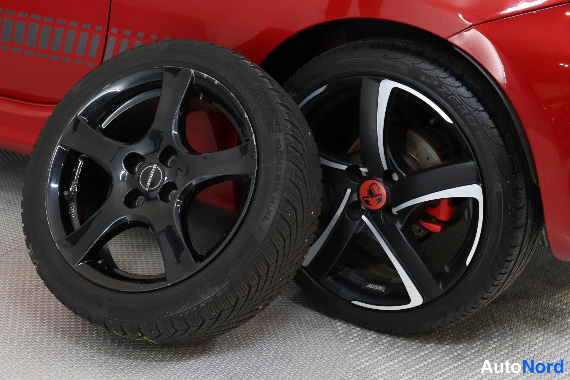 Yksityiskohta: Kaksi mustaa neljän peltin kevytmetallivannetta renkailla punaisen auton vieressä; toisessa Dunlop-merkkinappi ja 195/45 R 16 H XL -merkinnät, toisessa Abarth-k