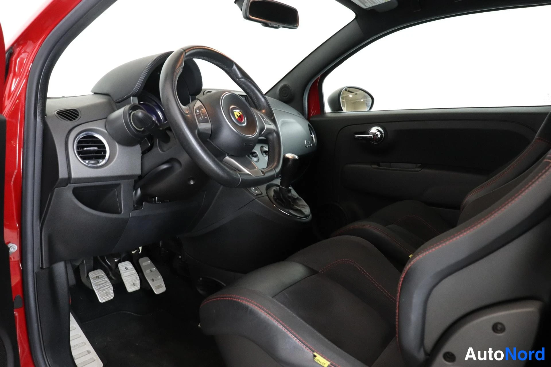 Ohjaamo: Interior shot showing the driver's area. The steering wheel features an Abarth badge at its center and silver multifunction buttons. Below, three metal pedals (