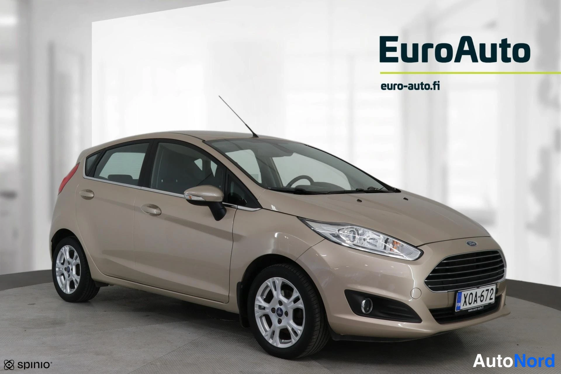 Etuviisto, nokka oikealle: Kultaharmaa Ford Fiesta -mallinen autotiehdasvalokuva. Auton edessä näkyy takapyörä ja keraamiset valot.