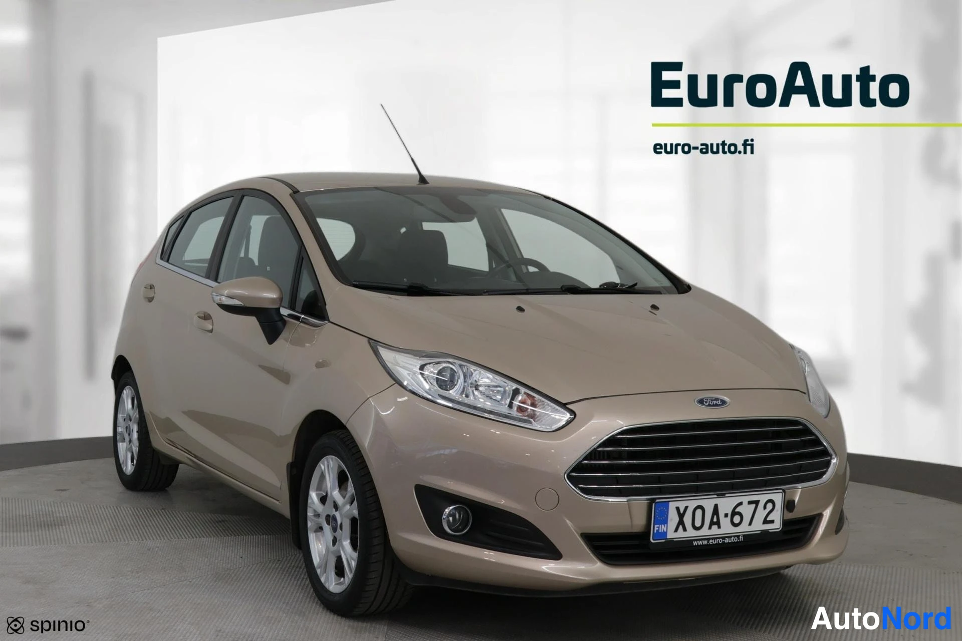 Edestä: Kultaharmaa Ford Fiesta -mallin etu- ja sivusäde. Alumiinirenkaat näkyvät.