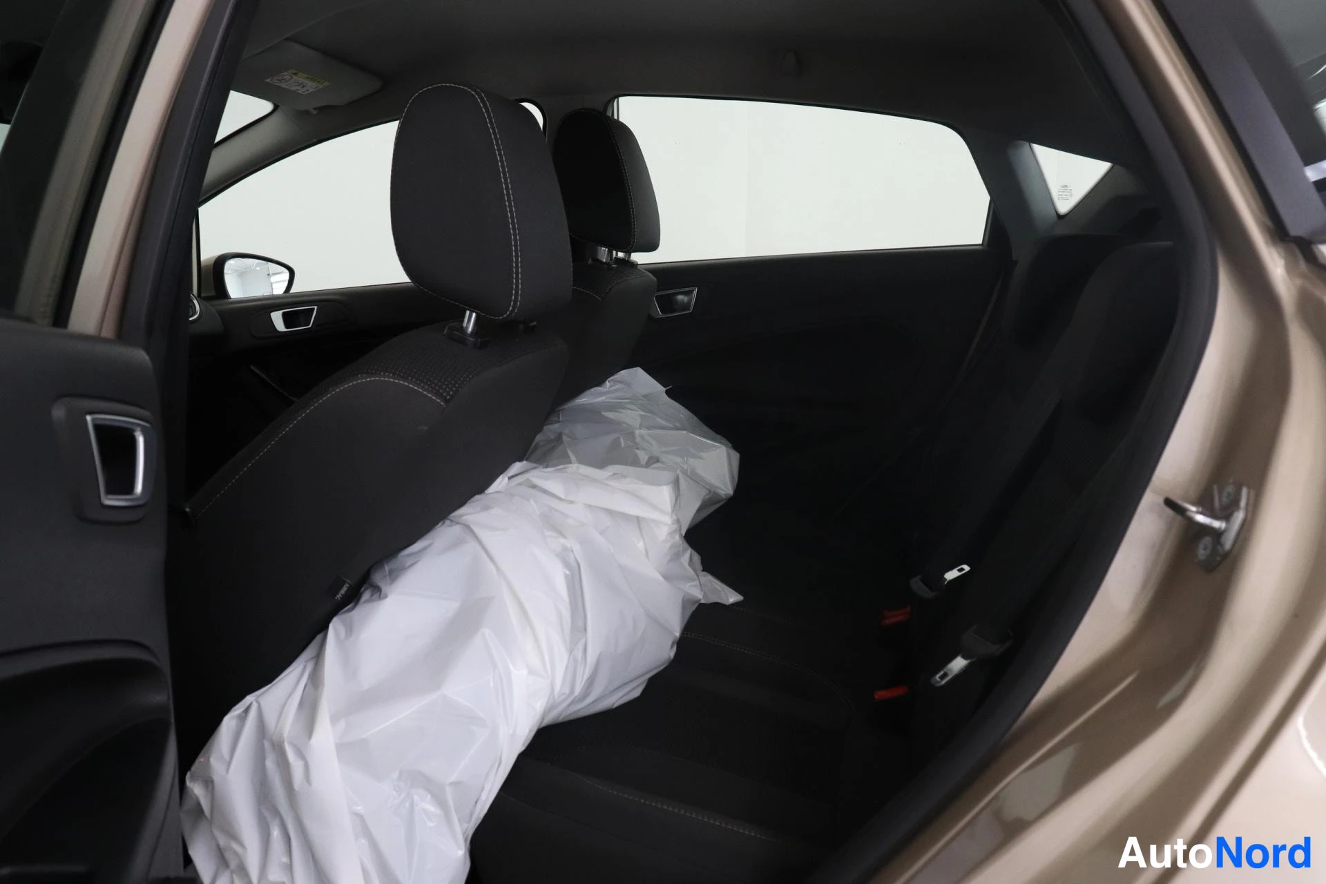 Takaistuimet: Rear passenger compartment view through open door showing two rear seats upholstered in dark fabric with contrasting stitching. White protective plastic wrappin