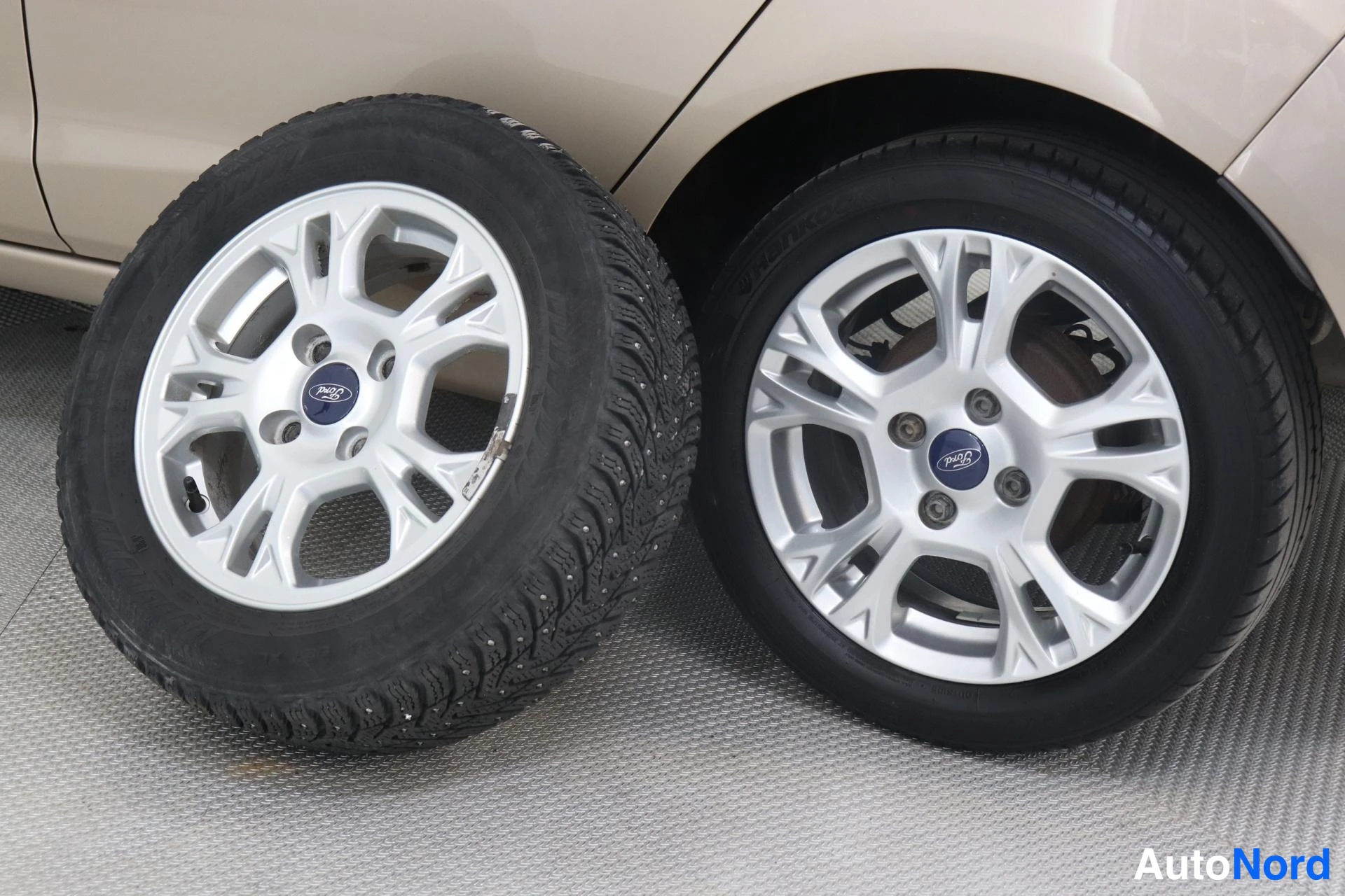 Sivulta: Kaksi Ford-alumiinivannetta: lattialla seisova rengas nastarenkaalla ja autossa kiinni oleva rengas, taustalla auton vaaleanbeige kylkipaneeli.