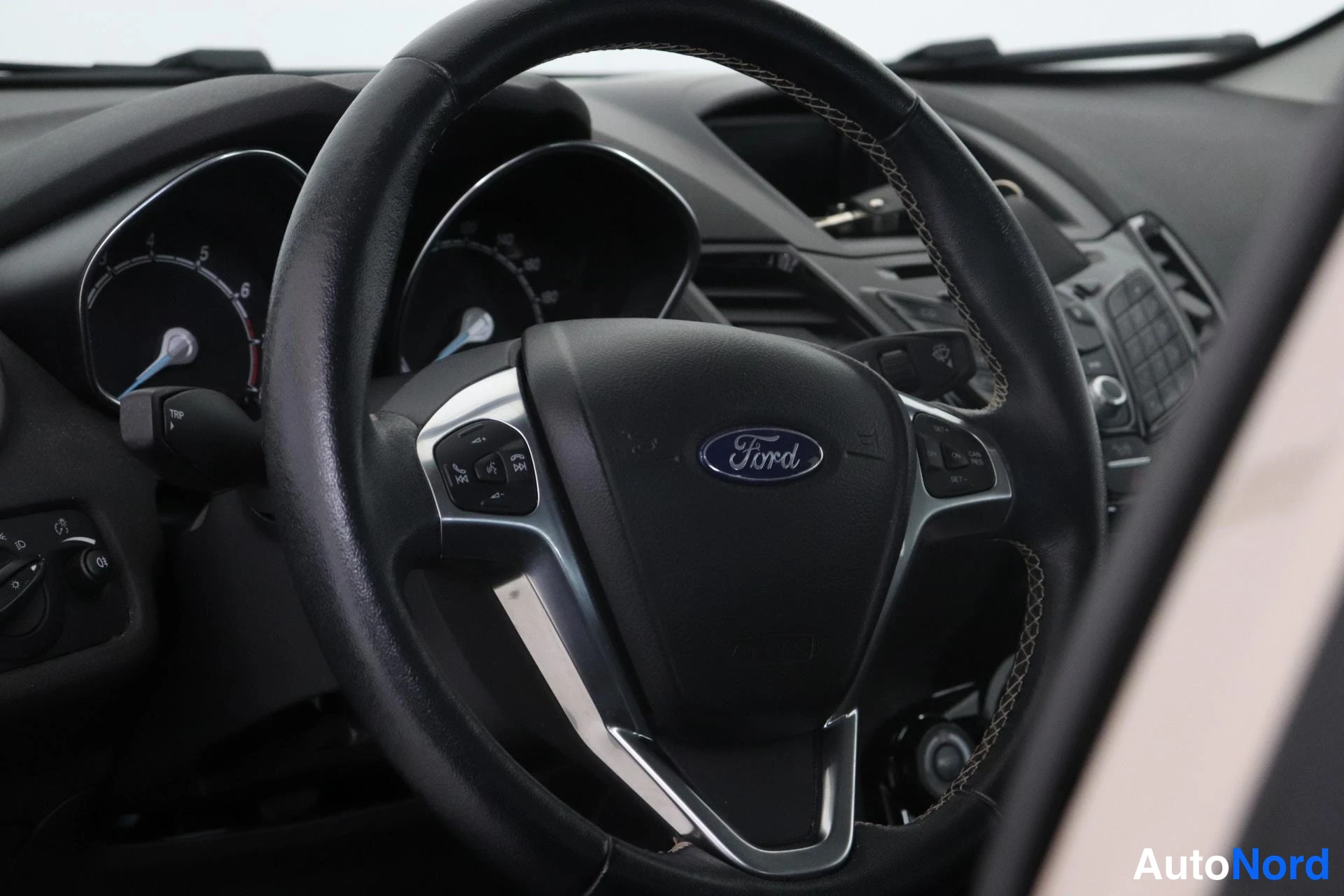 Edestä: Ford-merkkinen ohjauspyörä monitoimipainikkeineen lähikuvassa; taustalla näkyvät mittaristo sekä kojelaudan keskuskonsolin säätimet ja radio.