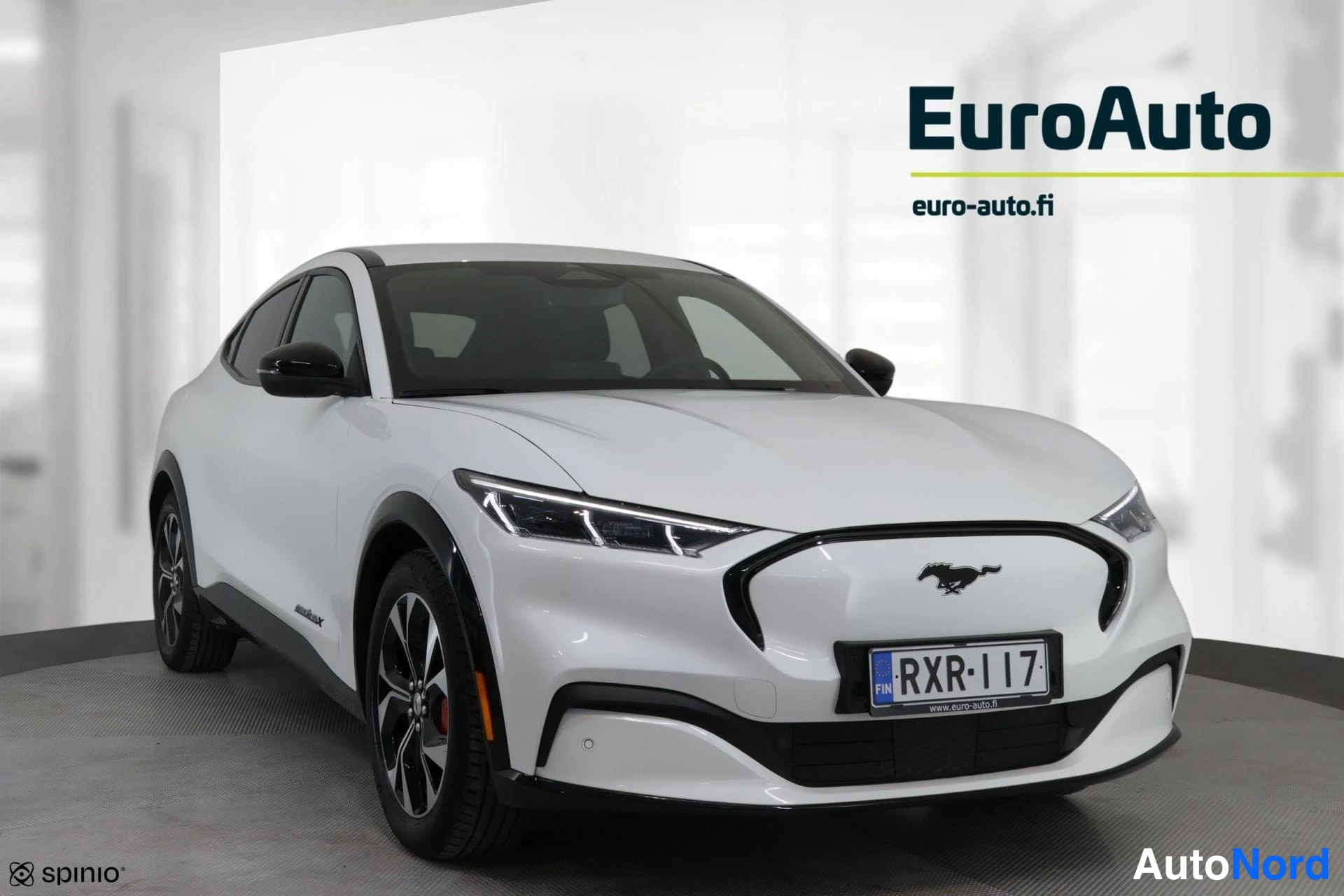 Etuviisto, nokka oikealle: Valkoinen Ford Mustang Mach-E etuviistokuva, keula kuvan oikealla; Mustang-merkki keulassa ja rekisteritunnus.