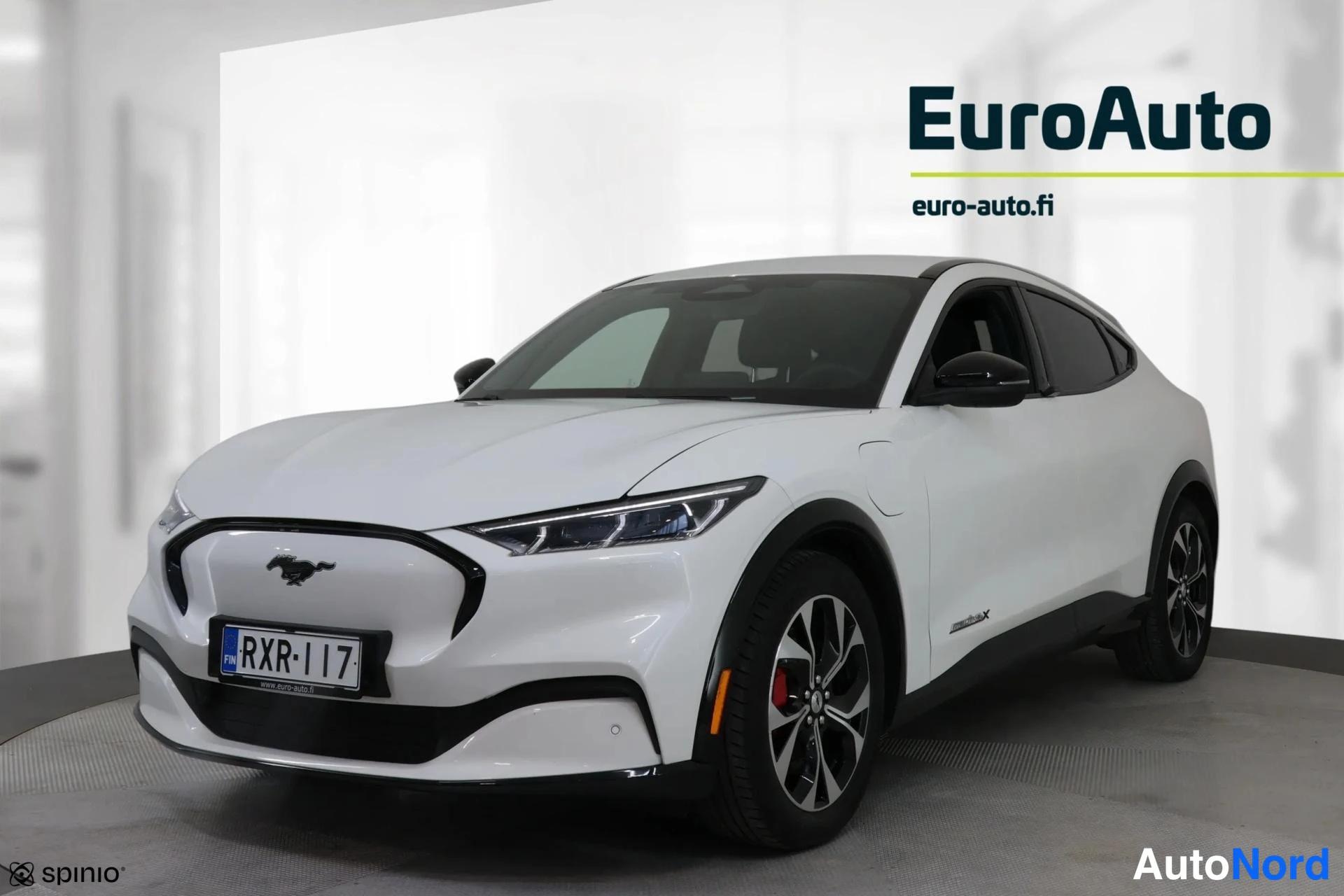 Etuviisto, nokka vasemmalle: Valkoinen Ford Mustang Mach-E etuviistokuva, keula vasemmalla; mustat kiiltavat hustut, hopeavannehitut ja rekisterikilpi; EuroAuto-logo taustalla.