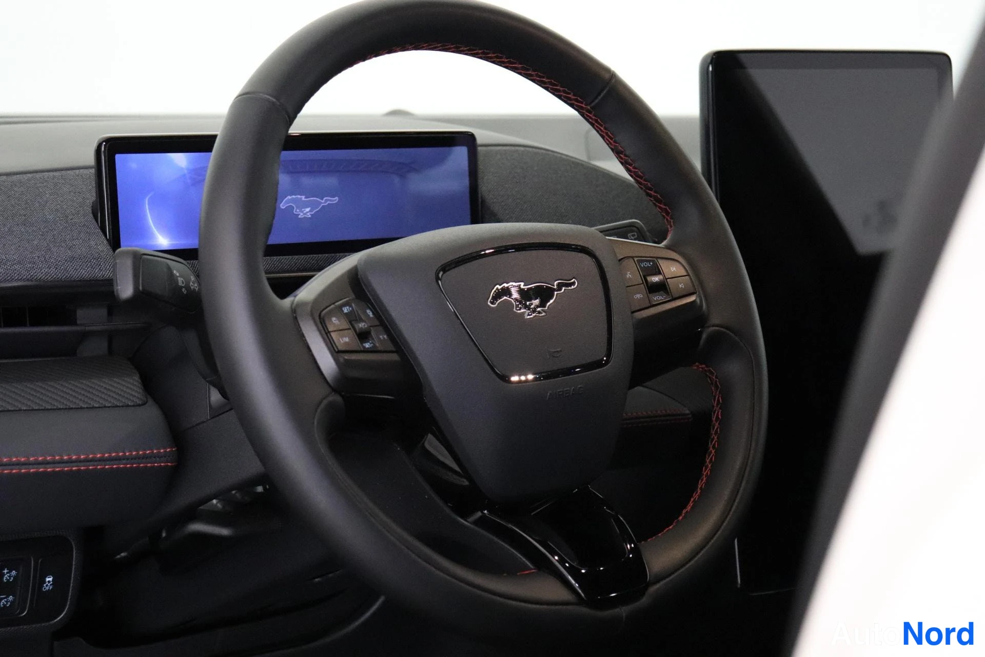 Muu: musta kolmivaiheinen ohjauspyörä, jossa punainen ommeltu reunustus ja Ford Mustang -logo. Takana on sinisellä näytöllä varustettu instrumenttilauta.