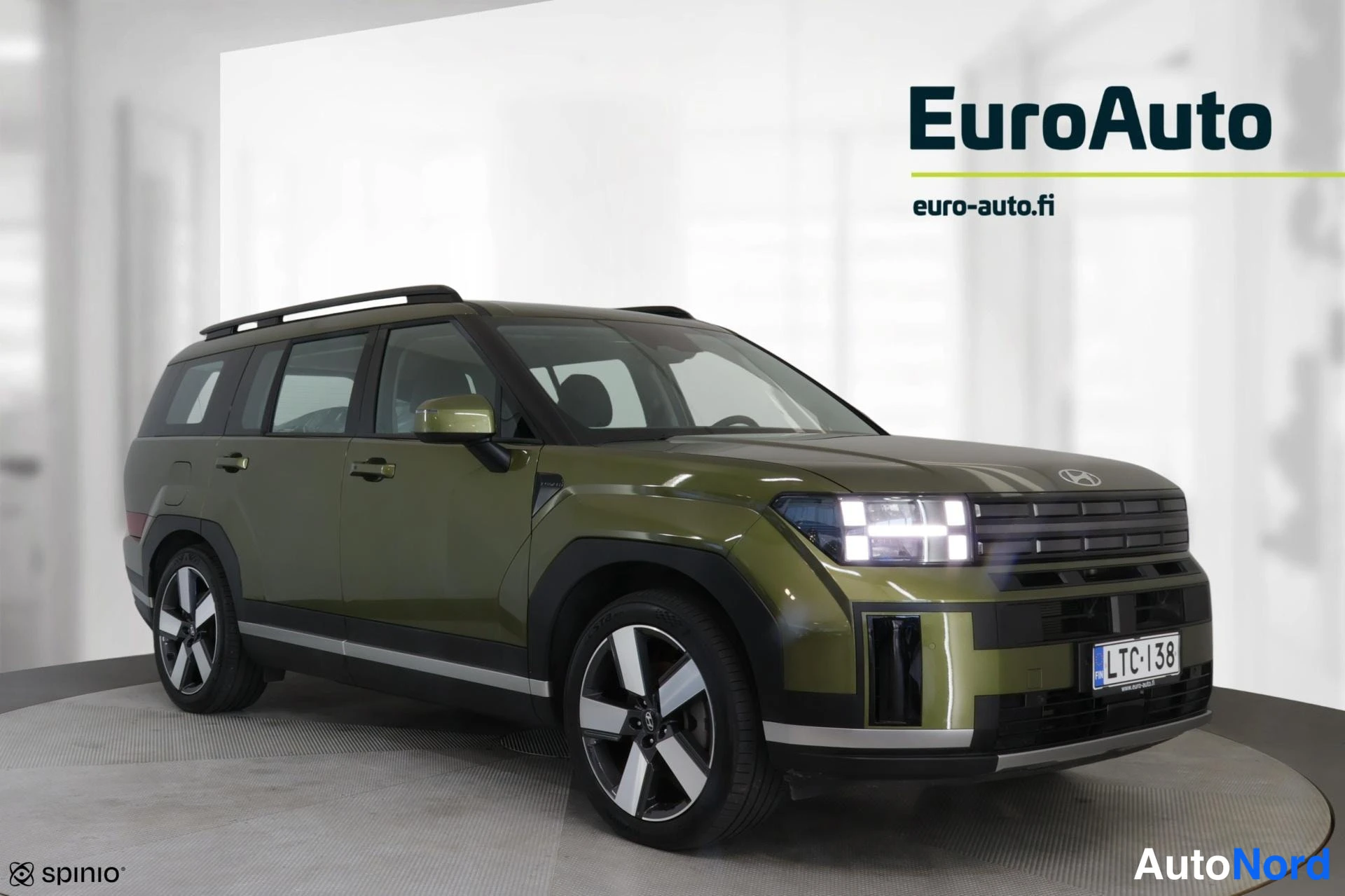Etuviisto, nokka oikealle: Etuvino kuva oliivinvihreästä Hyundai Santa Fe -maasturista, keula oikealla; kattokaiteet, ohjausvalot ja rekisterikilpi erottuvat studiotaustaa vasten.