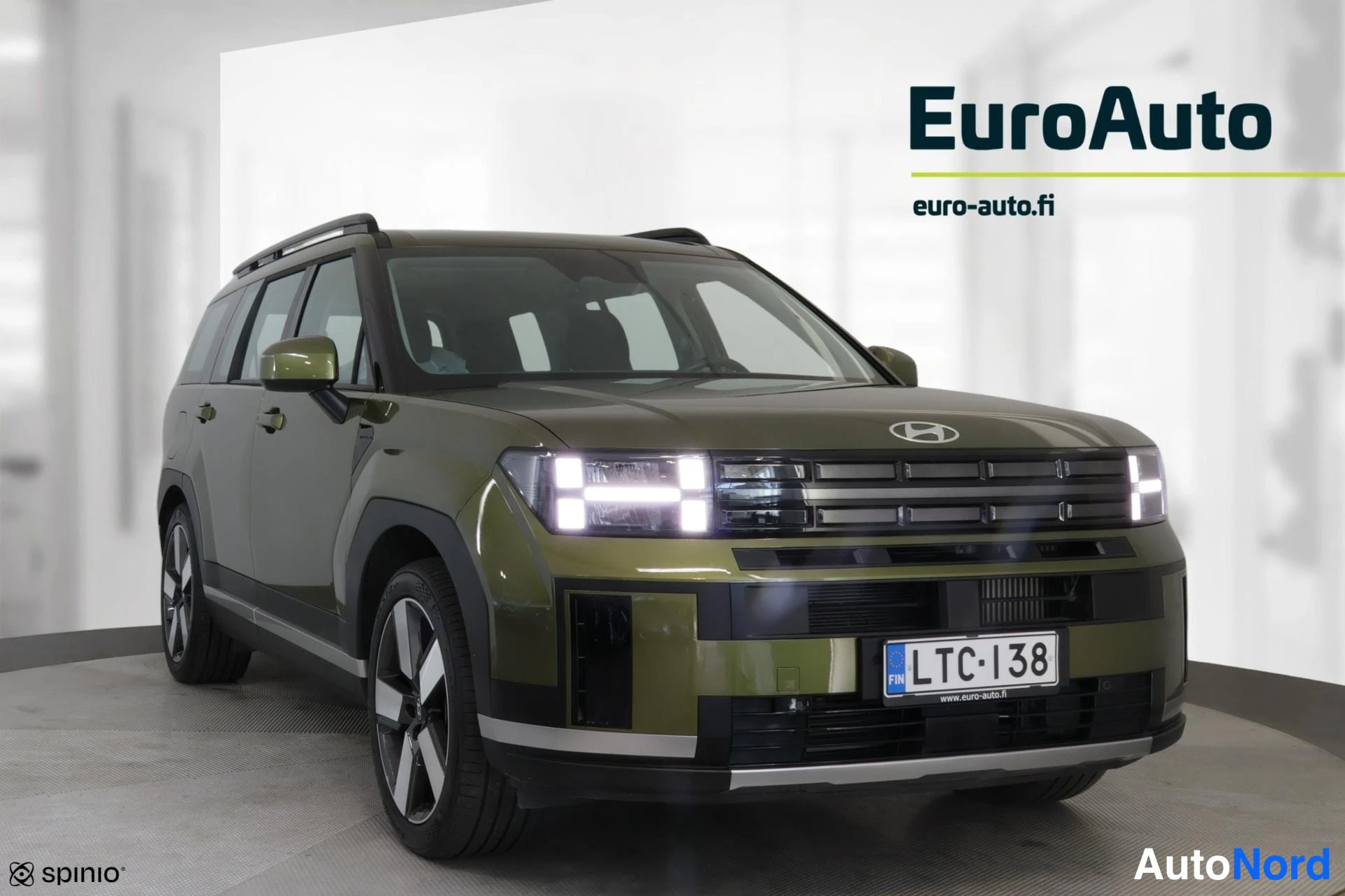 Etuviisto, nokka oikealle: Etuvino lähikuva oliivinvihreästä Hyundai Santa Fe -maasturista, keula oikealla; verkkomainen etusäleikkö, LED-ohjausvalot ja kylki näkyvät, rekisterikil