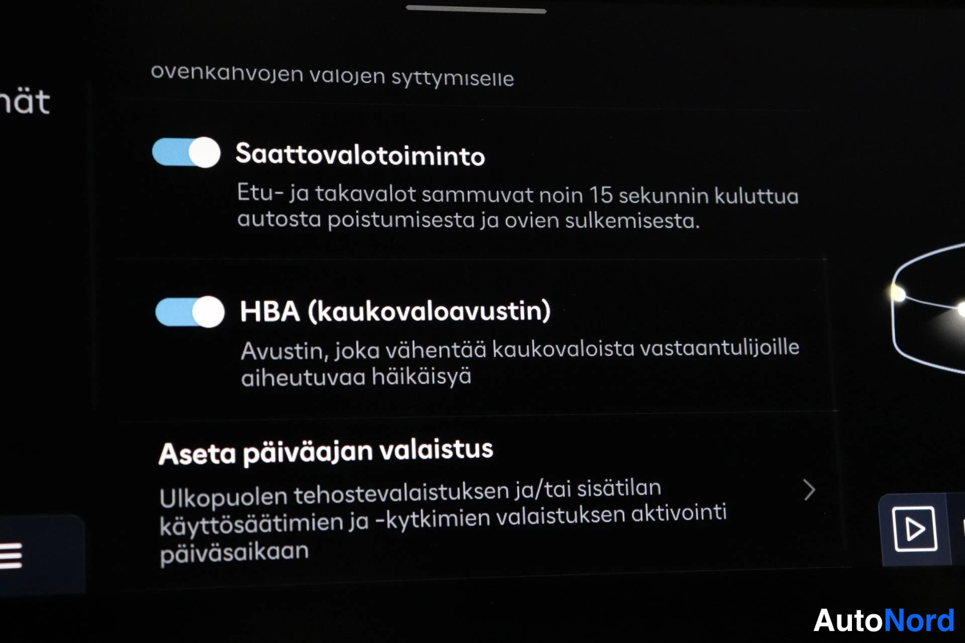 Yksityiskohta: Infotainment-näytön kuva: valikko jossa Saattovalotoiminto ja HBA (kaukovaloavustin) päällä sekä päiväajan valaistuksen asetus.