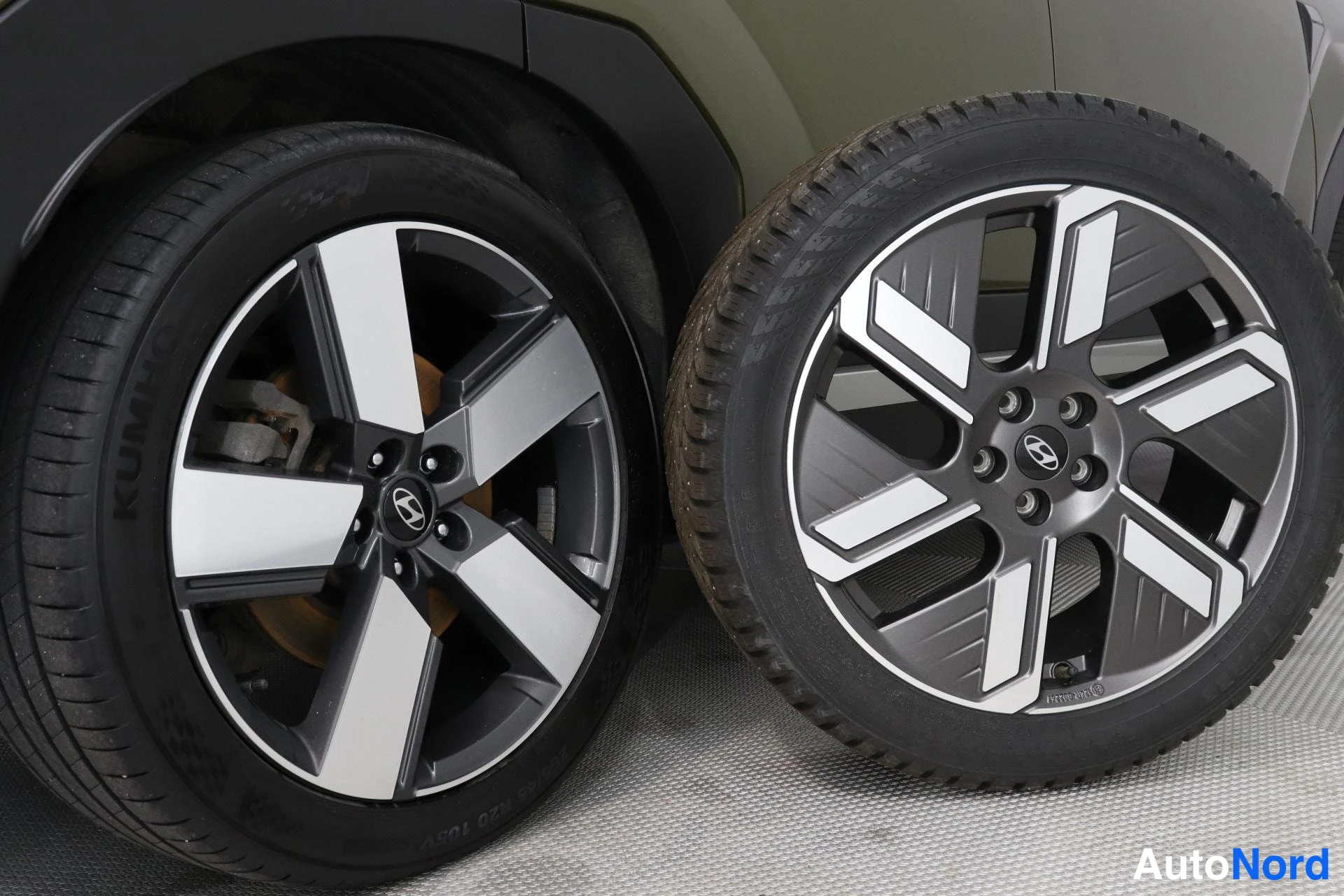 Muu: Kaksi viisipeäistä kevytmetallivannetta nokkarenkailla Hyundai-keskuskoristeella: toinen rengas kiinnitettynä autoon, toinen irrallisena lattialla sen vieressä.