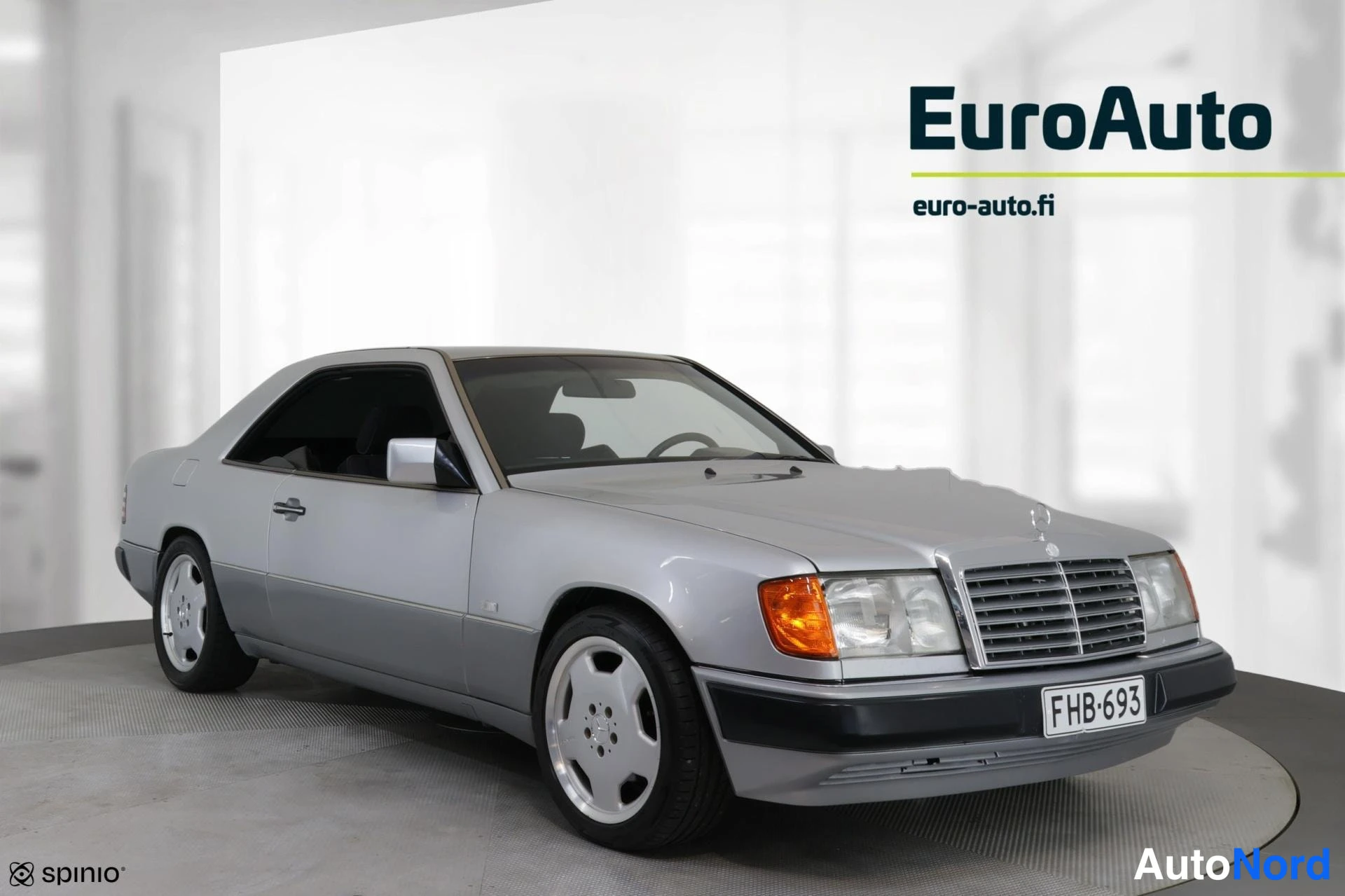 Etuviisto, nokka vasemmalle: Silver Mercedes-Benz coupe shown from the front-left side in a white showroom. The vehicle's body panels and doors are silver. Visible equipment includ