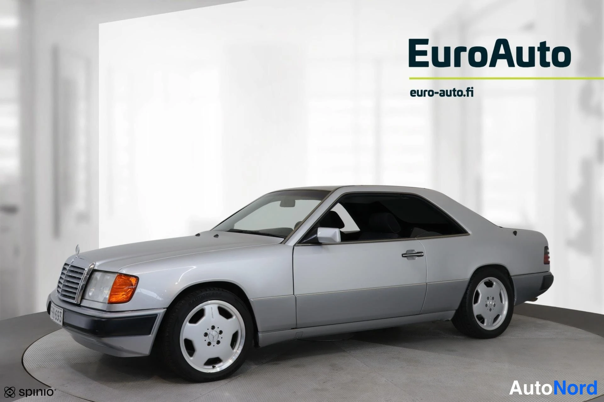 Etuviisto, nokka vasemmalle: Hopeinen Mercedes-Benz W124 -coupé vinosti edestä vasemmalta valkoisessa studiossa