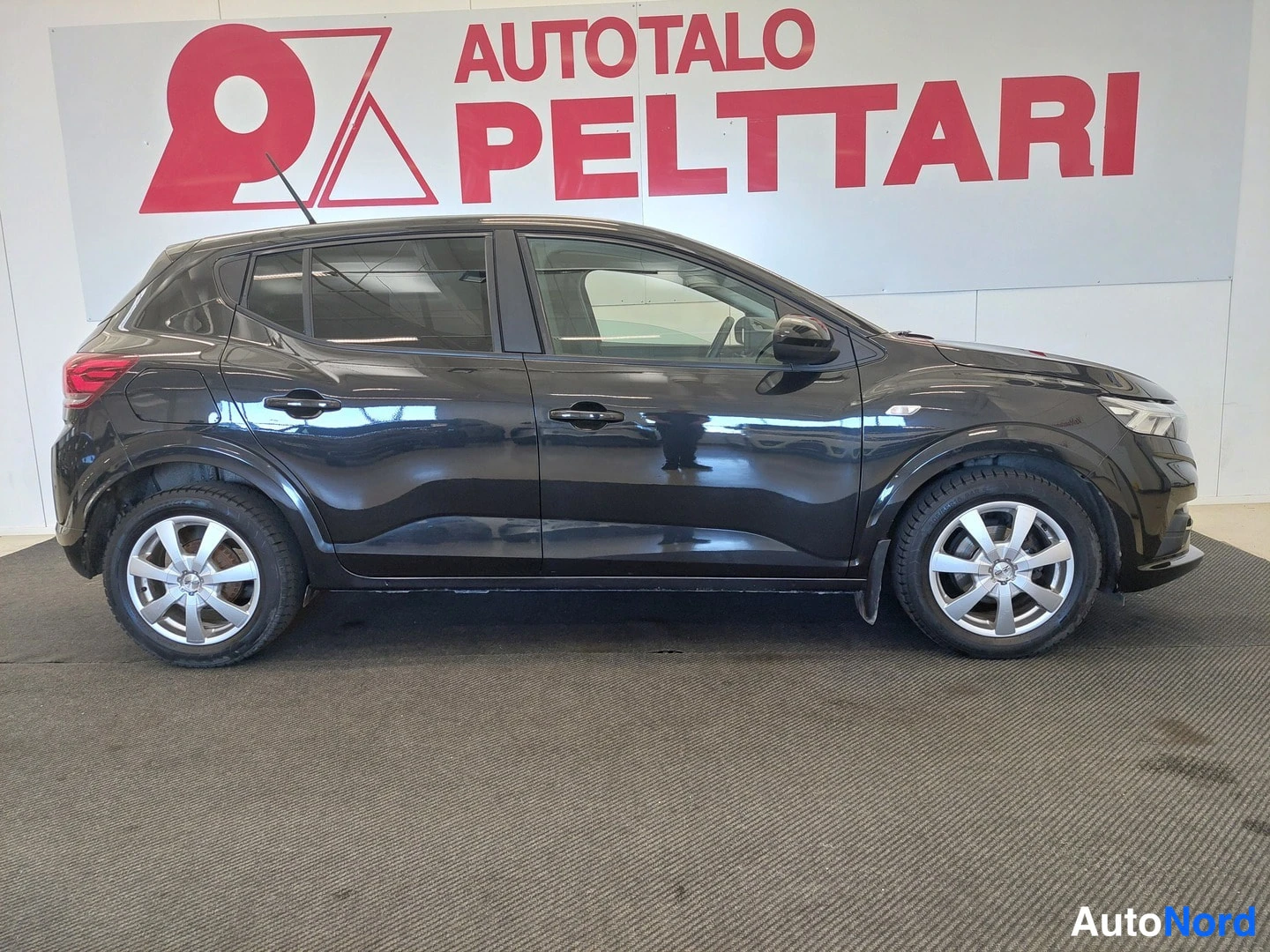 Etuviisto, nokka oikealle: Mustan hatchbackin kylkikuva sisätiloissa, keula oikealla, taustalla Autotalo Pelttari -mainostaulu.