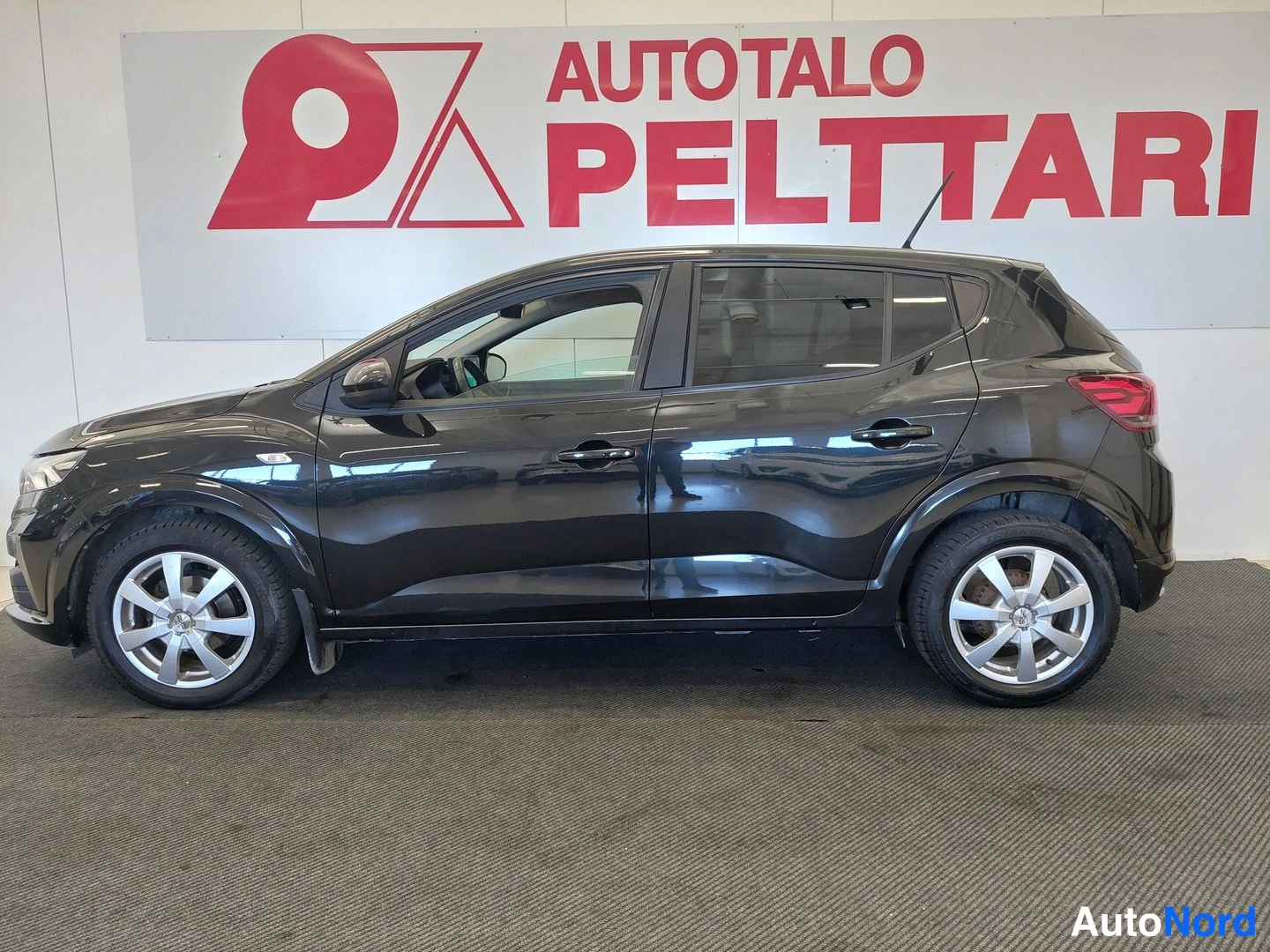 Sivulta: Musta Dacia Sandero auto seisomassa sisällä sivukuvassa. Auton renkaat ovat hopeanvärisiä kevytmetallivanteita. Taustalla punainen ja valkoinen seinä mainoksell