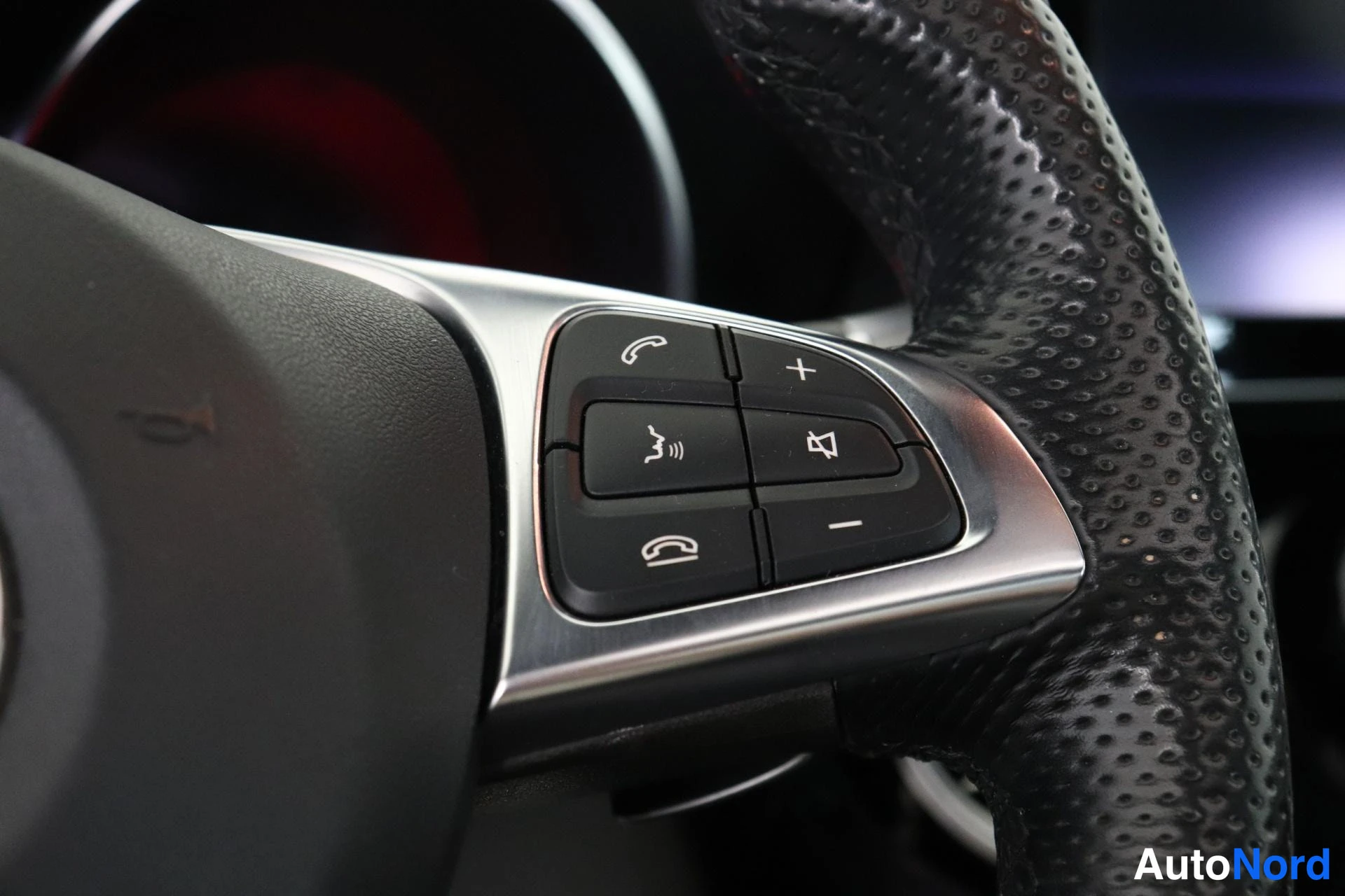 Yksityiskohta: Close-up of the right side of the steering wheel with audio and phone controls: call answer/end buttons, plus/minus volume knobs, and mute button on black perfo