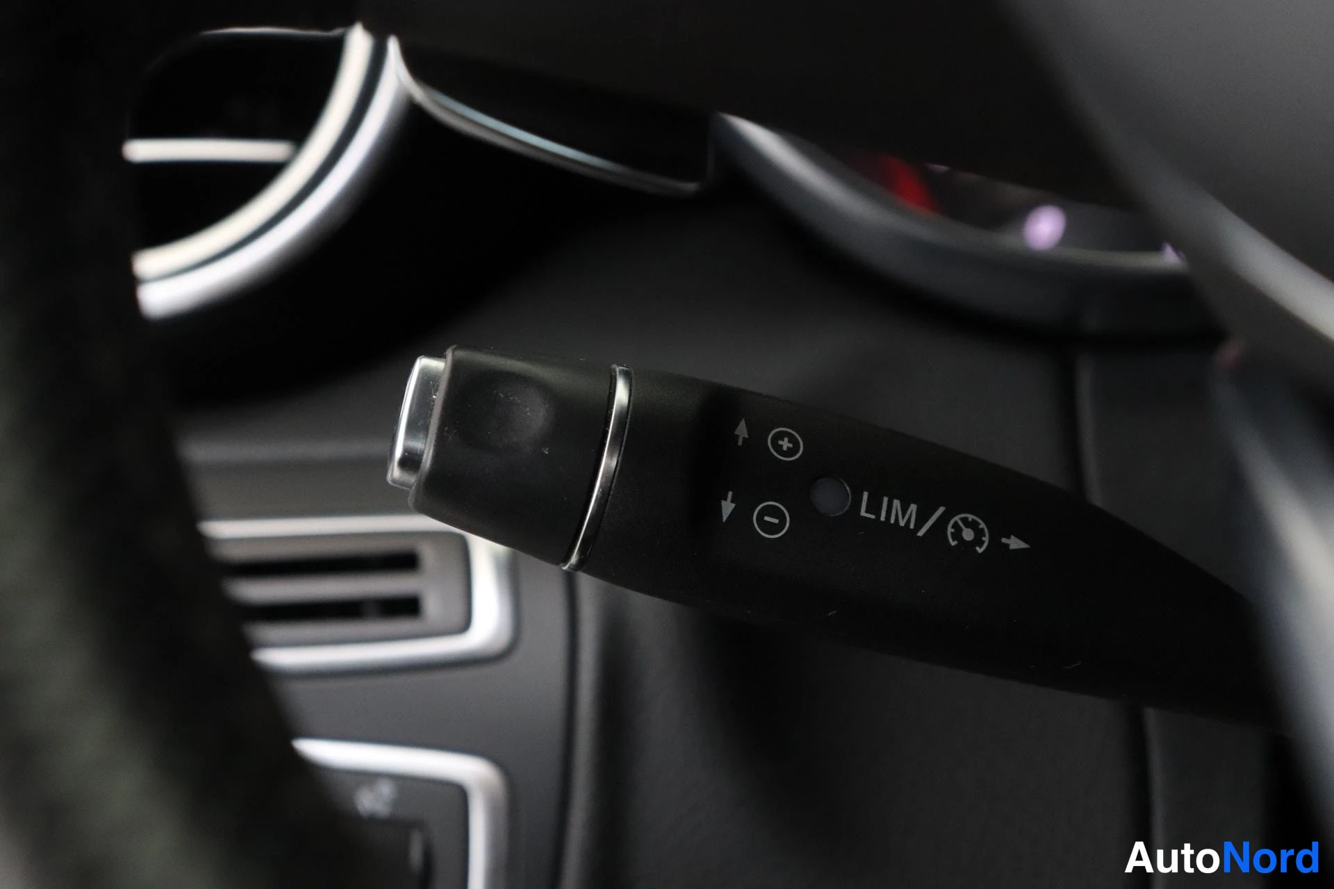 Yksityiskohta: Stalk mounted behind the steering wheel labeled LIM/ for speed limiter (with + / - adjustment symbols), set against dark dashboard trim near circular air vent.