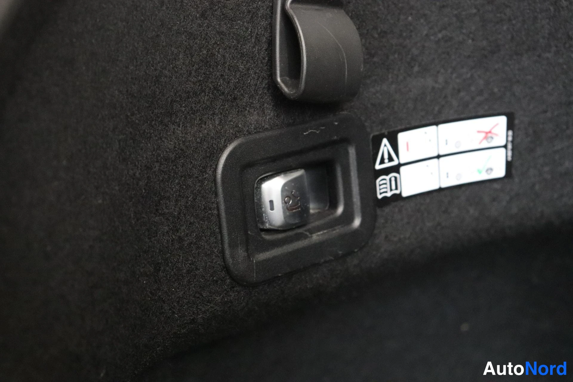 Tavaratila: Lähikuva takakamon sisäpinnalta: ISOFIX-lukitus (metallinen kiinnike), varoitustarra symbolineen ja musta kangas.