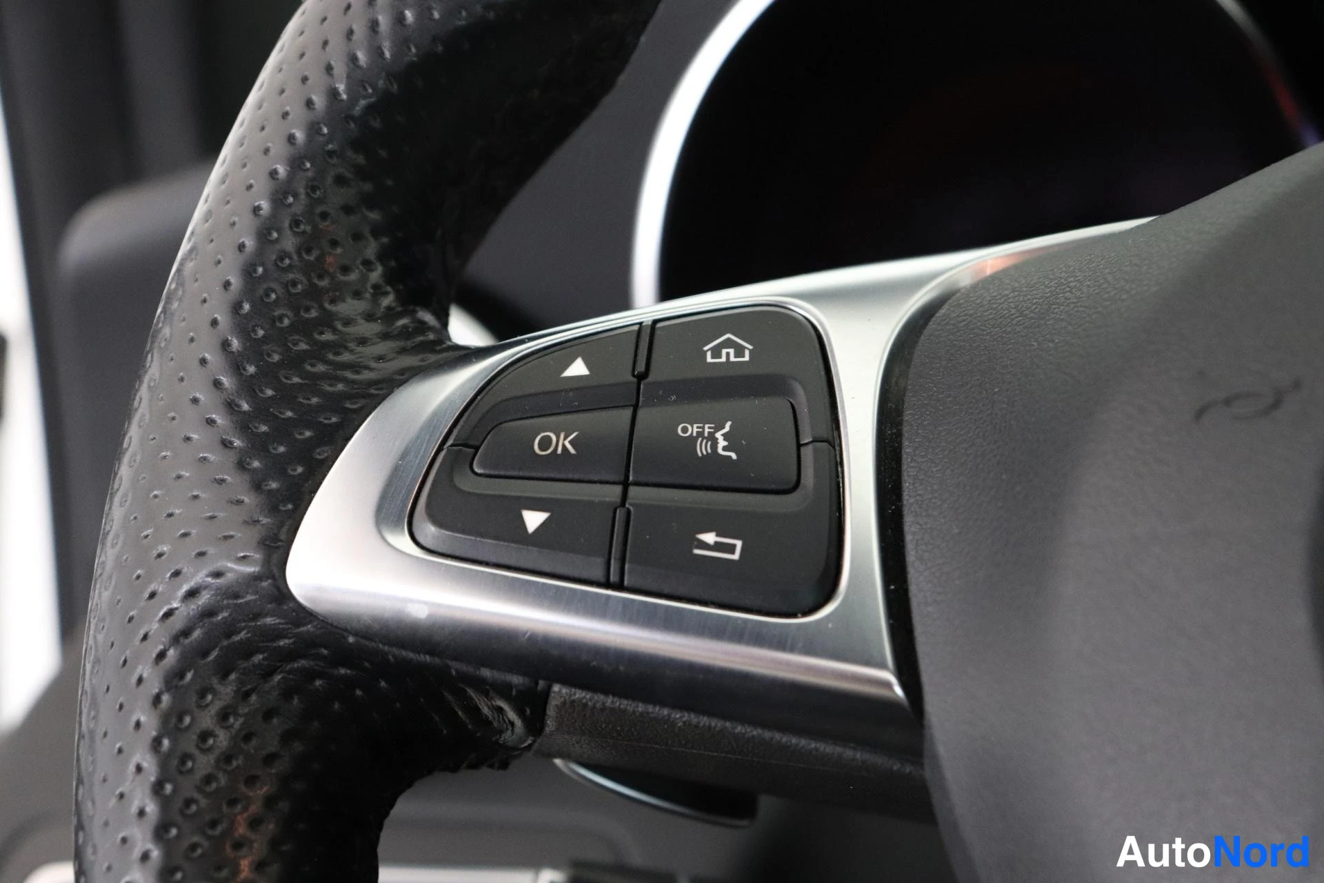 Yksityiskohta: Close-up of the left side of the steering wheel featuring menu navigation controls, including directional arrows, an OK button, a home icon, and a cruise contro