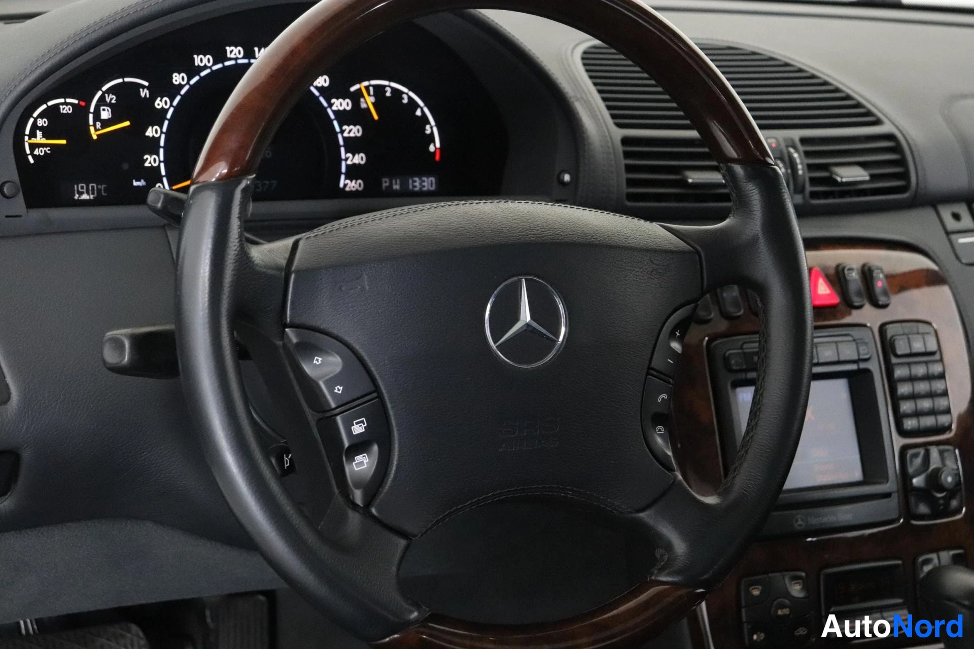 Sivulta: Kuljettajan näkymä: puupintainen nahkainen Mercedes-ohjauspyörä, mittaristo, ja puupaneloitu keskikonsoli näytöllä, painikkeilla ja varoituskolmiopainikkeella.