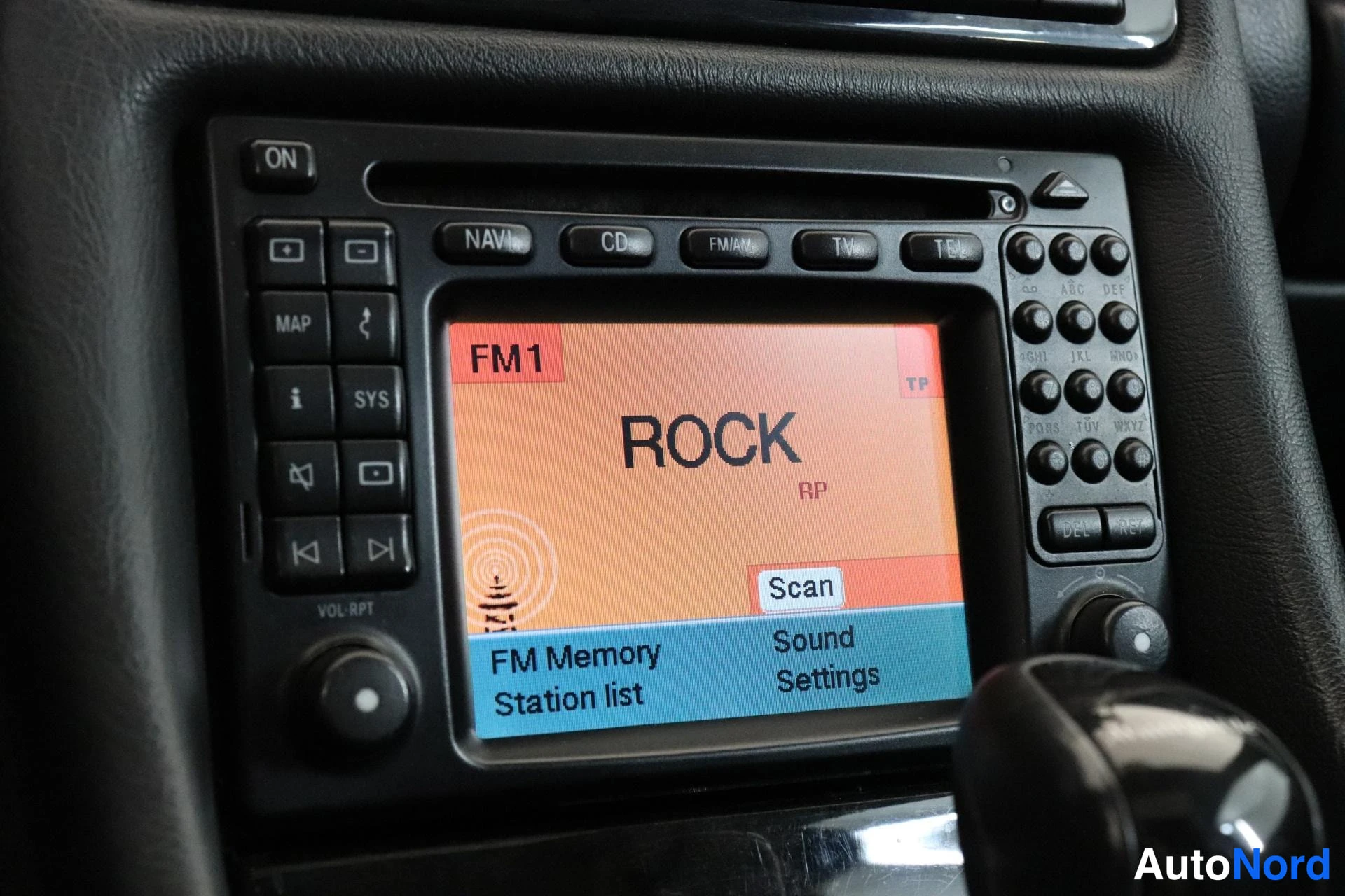 Yksityiskohta: Kuva auton navigaatiojärjestelmän kosketusnäytöstä ja sen sivulla olevista ohjauspainikkeista. Näytössä lukee FM1-radioasemalla rockmusiikkia ja sen alla ovat v