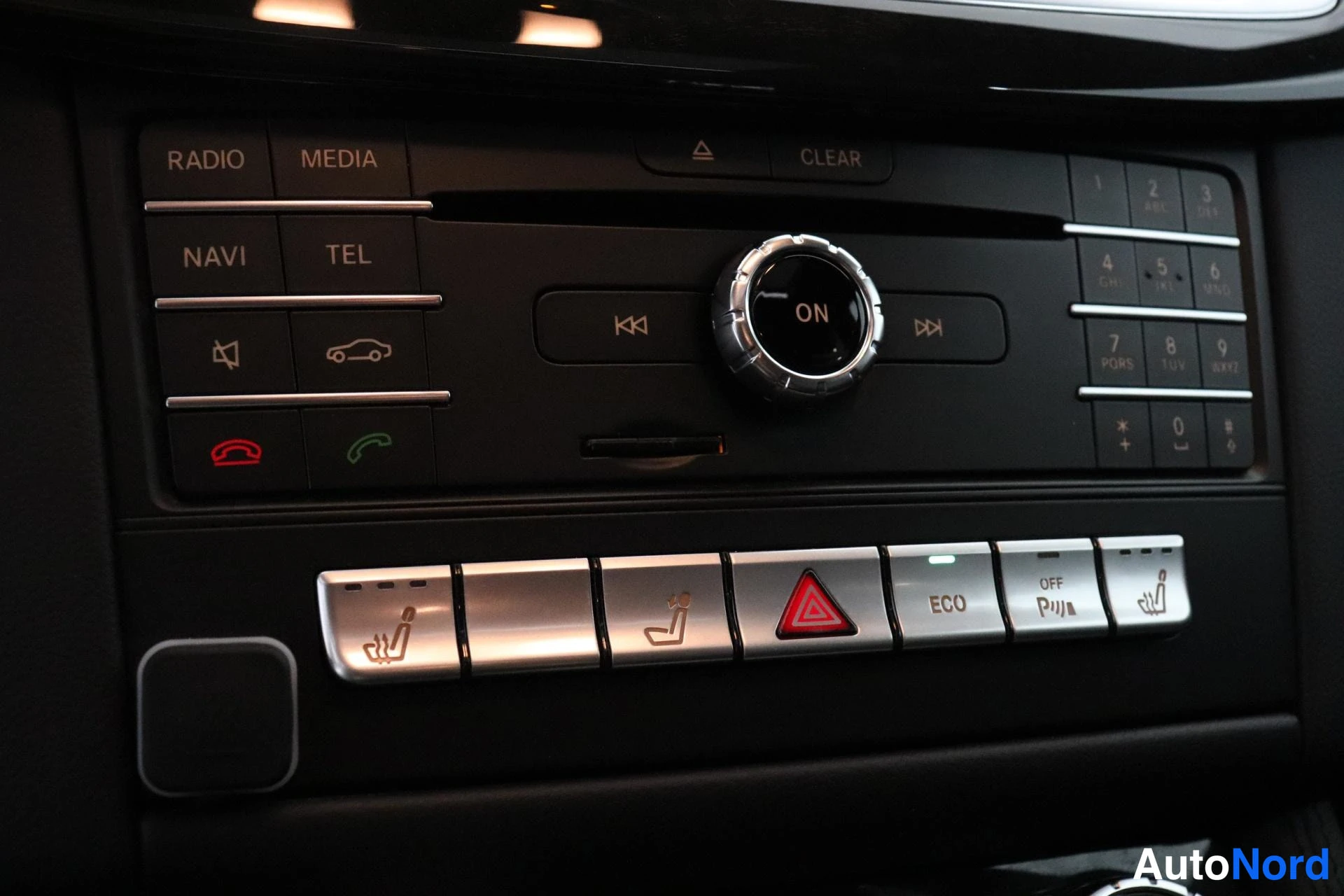 Yksityiskohta: Close-up of centre stack showing buttons for RADIO, MEDIA, NAVI and TEL. Below are silver-finished controls with heated seat icons, an ECO button and a traction