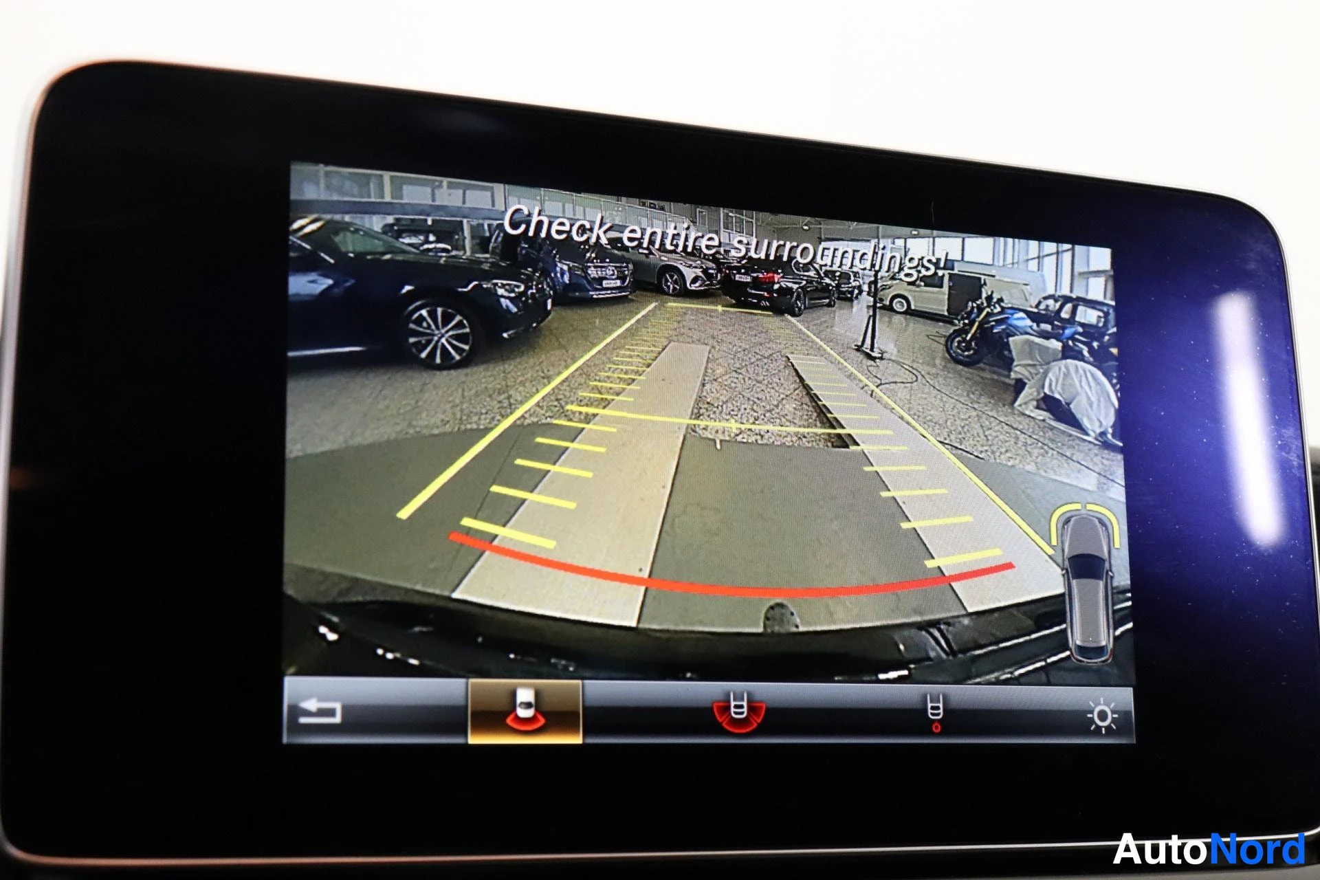 Ohjaamo: In-car display screen showing the rear-view camera feed with dynamic yellow and red guidance lines overlaying the parking area behind.