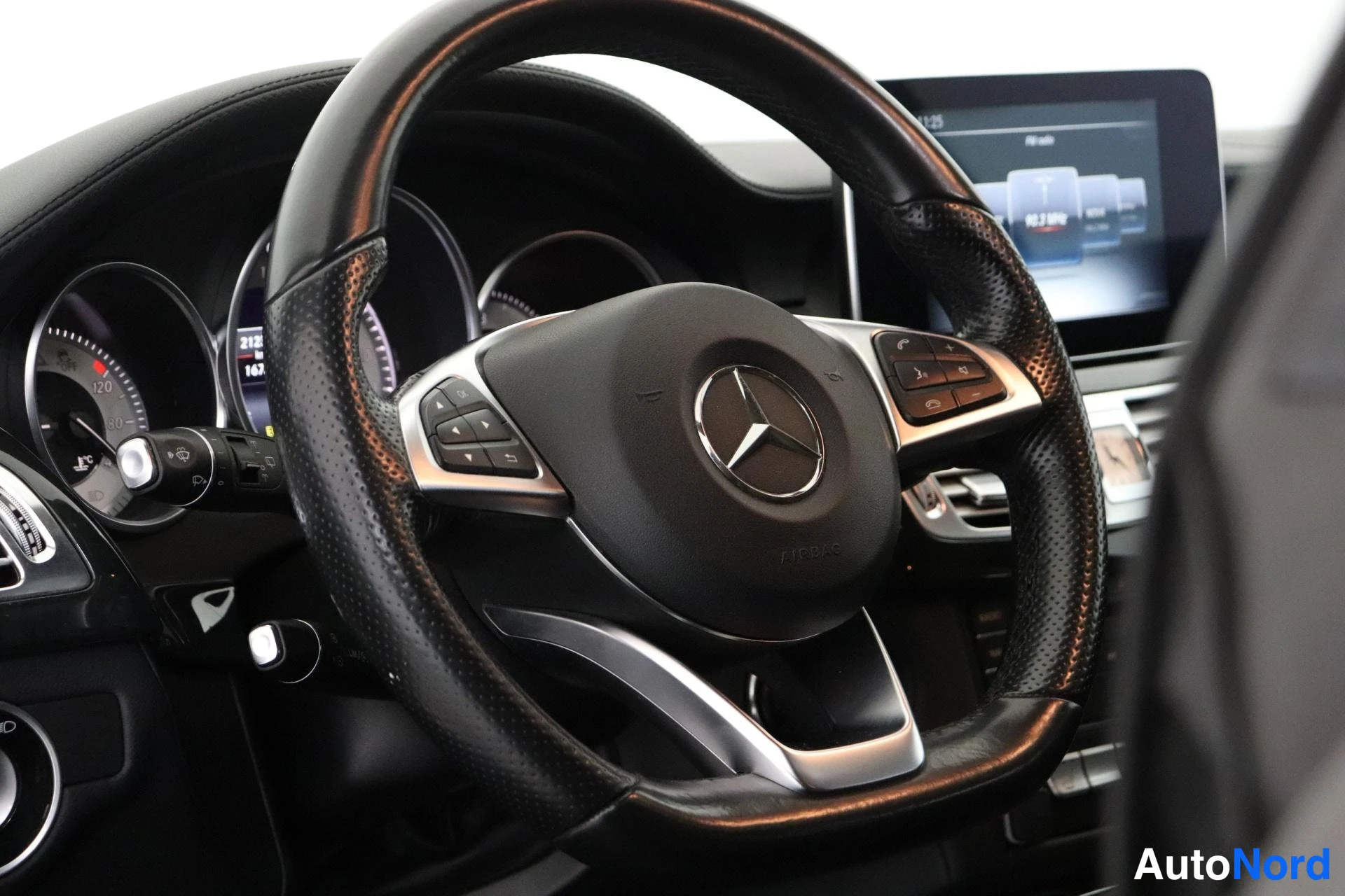 Sivulta: Ohjauspyörä Mercedes-tähdellä ja aukotetussa nahassa, mittaristo sekä keskinäyttö, jonka Radio-valikko ja kello näkyvät.