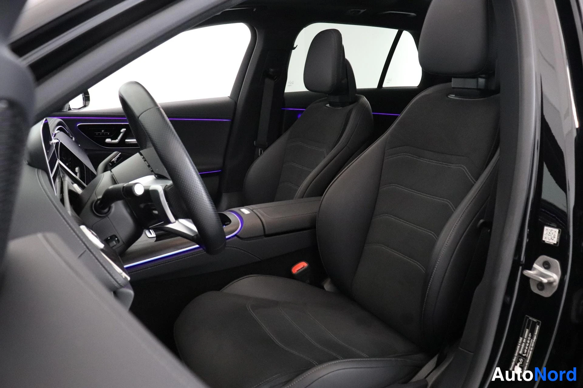 Ohjaamo: Interior view from the driver's side door showing two black front seats upholstered in perforated material. Blue ambient lighting strips run along the dashboard