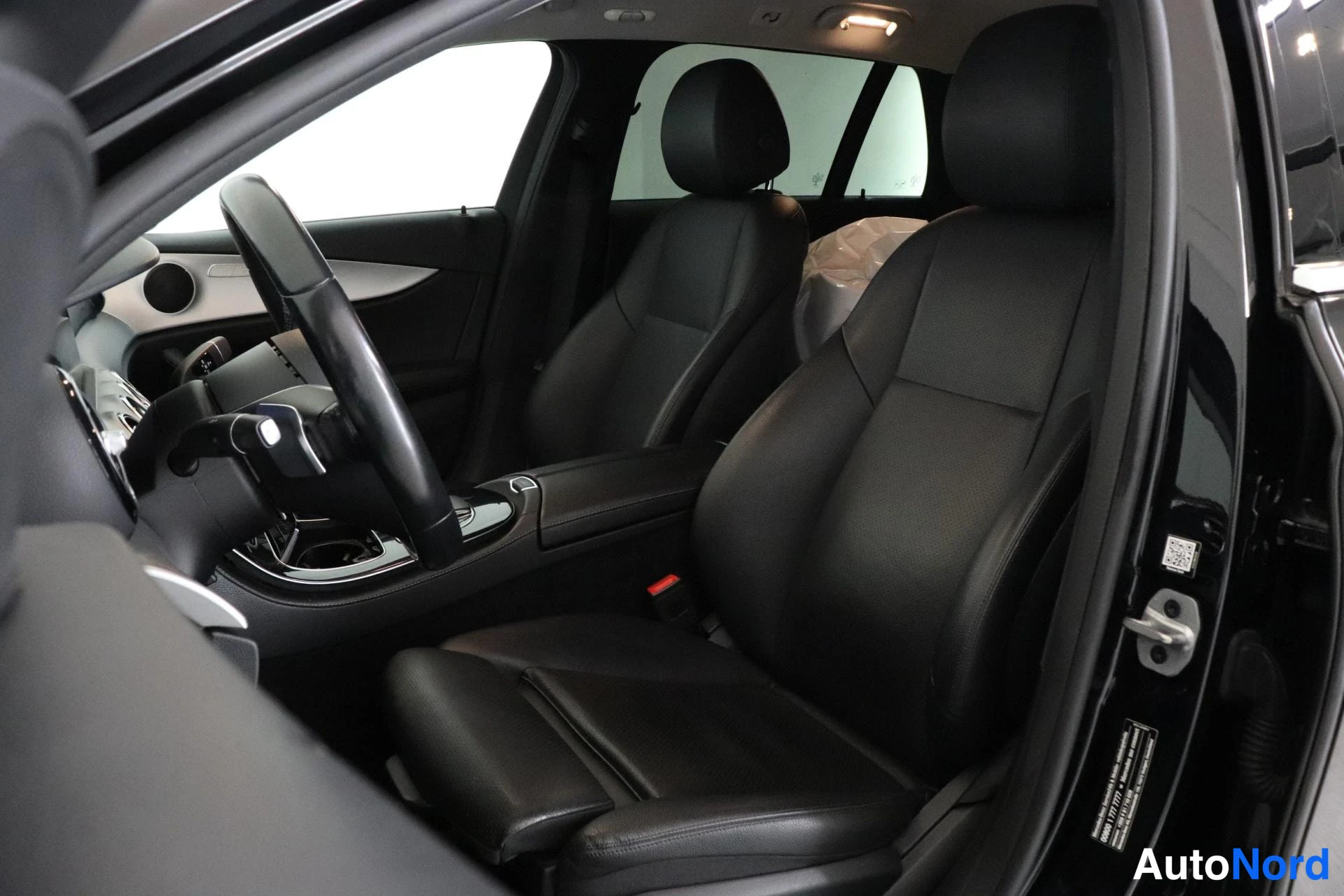 Sivulta: Interior view looking into the front cabin through the open driver's side door. Visible are black leather front seats, the steering wheel, dashboard vents, and