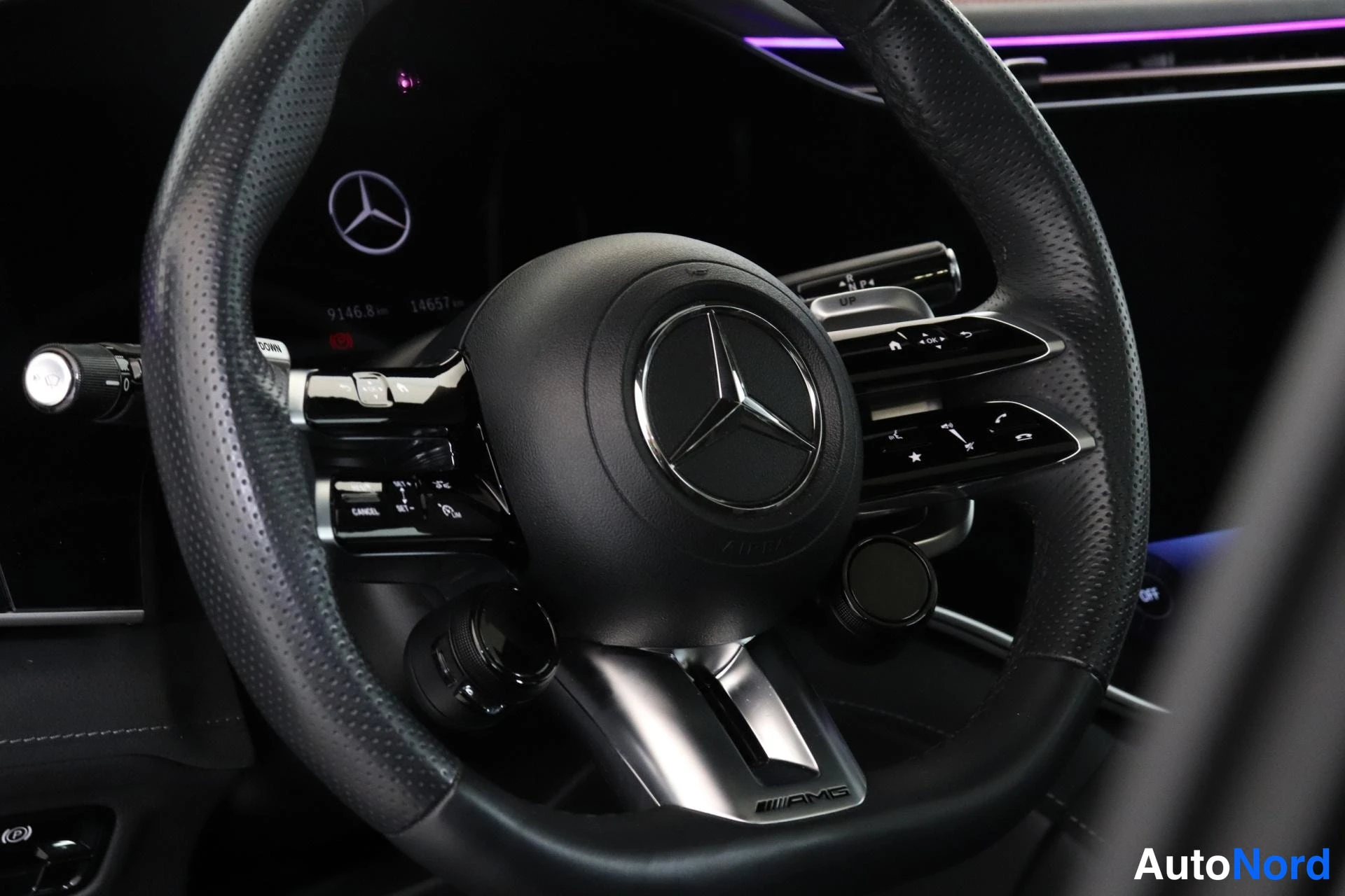 Yksityiskohta: Ohjauspyörän lähikuva. Sen keskellä on Mercedes-Benz-logo ja se on varustettu useilla hallintapainikkeilla. Ohjauspyörän alla on teksti AMG. Taustalla mittarist