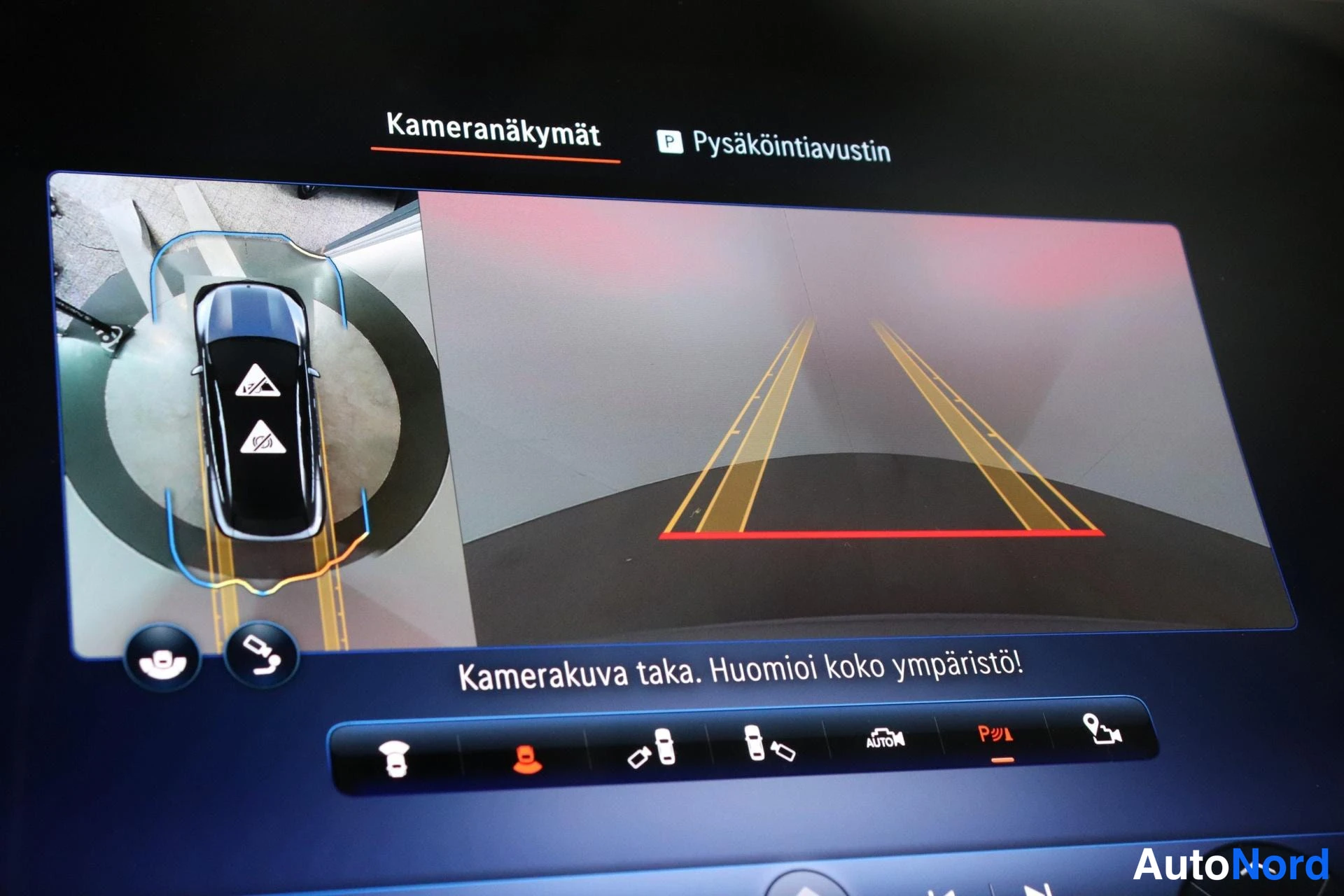 Yksityiskohta: Infotainment-näyttö lähikuvassa: kamera näkymät -valikko, ylhäältä kuvattu auto ja peruutuskameran kuva ohjelinjoineen; teksti 'Kamerakuva taka. Huomioi koko ym