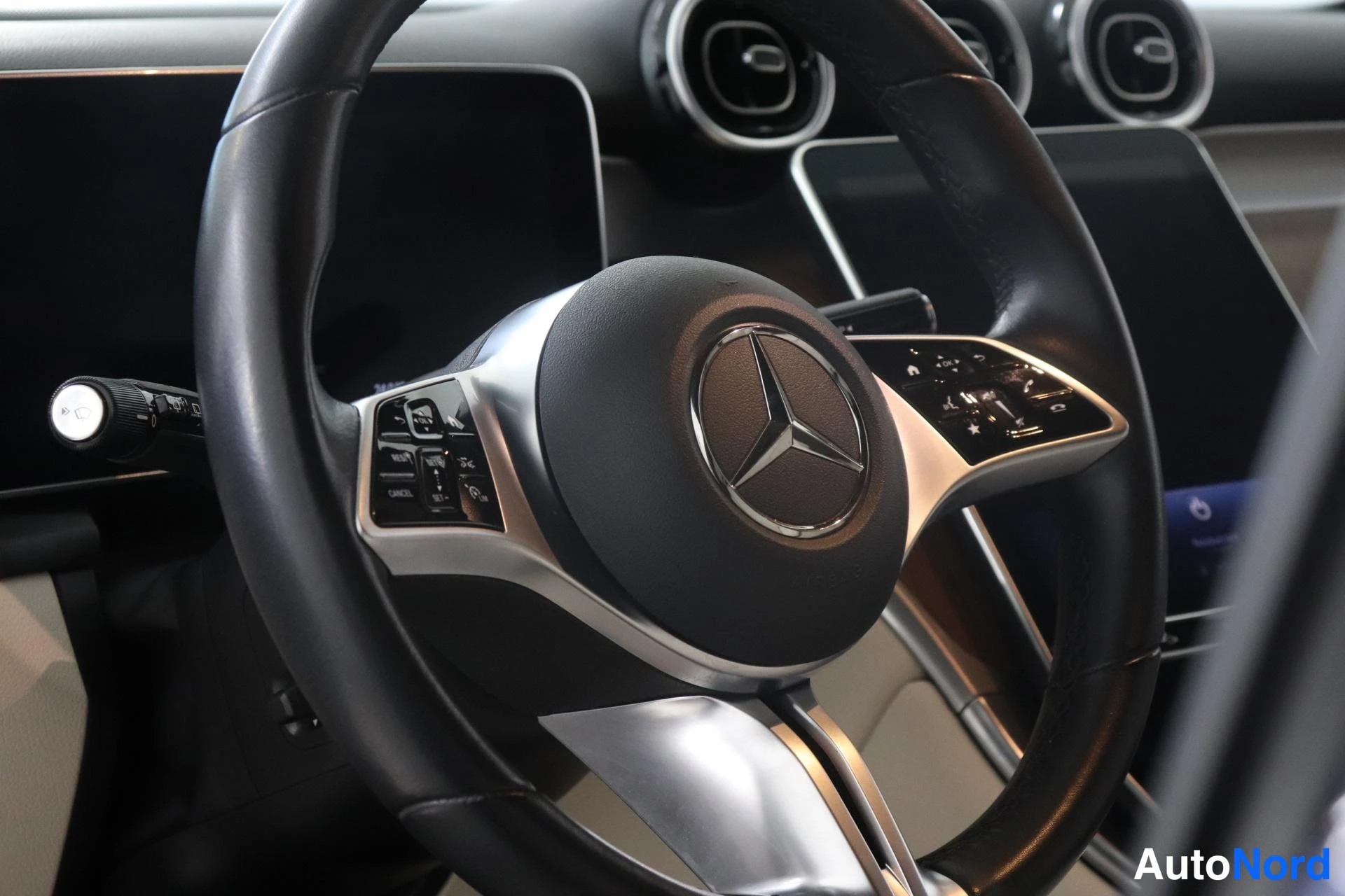 Etuistuimet: Close-up of a black leather-wrapped multifunction steering wheel with the Mercedes-Benz logo on its central hub. On the left spoke are buttons including cruise