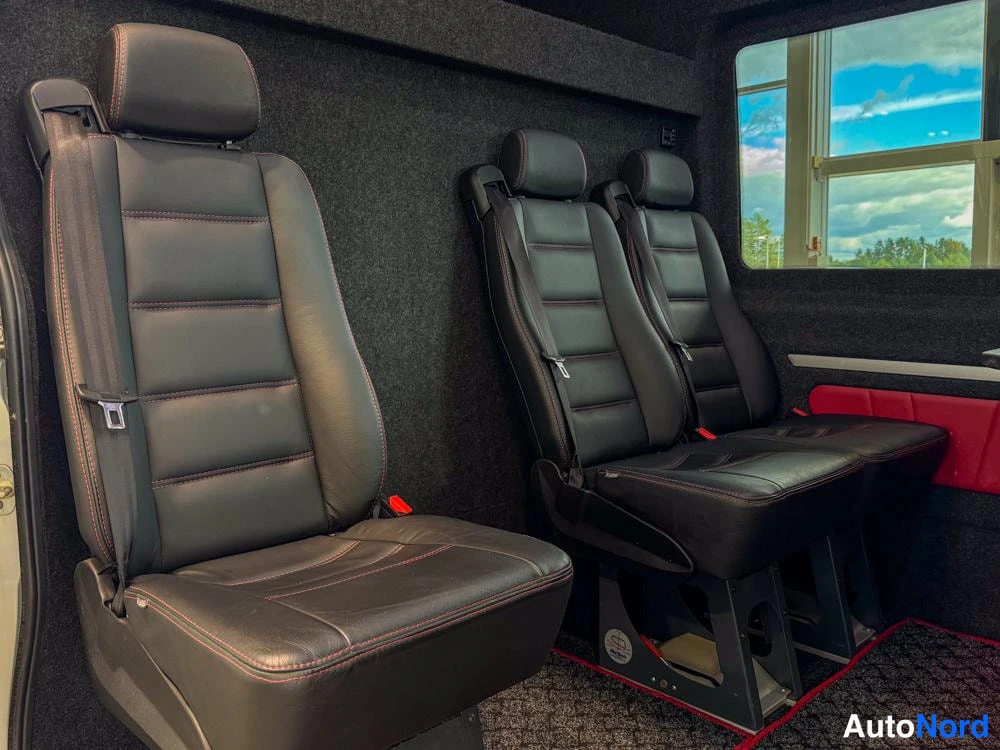 Ohjaamo: Close-up view of the black leather passenger seats inside the van, featuring red contrast stitching and integrated headrests. The seat mounting rails are visibl